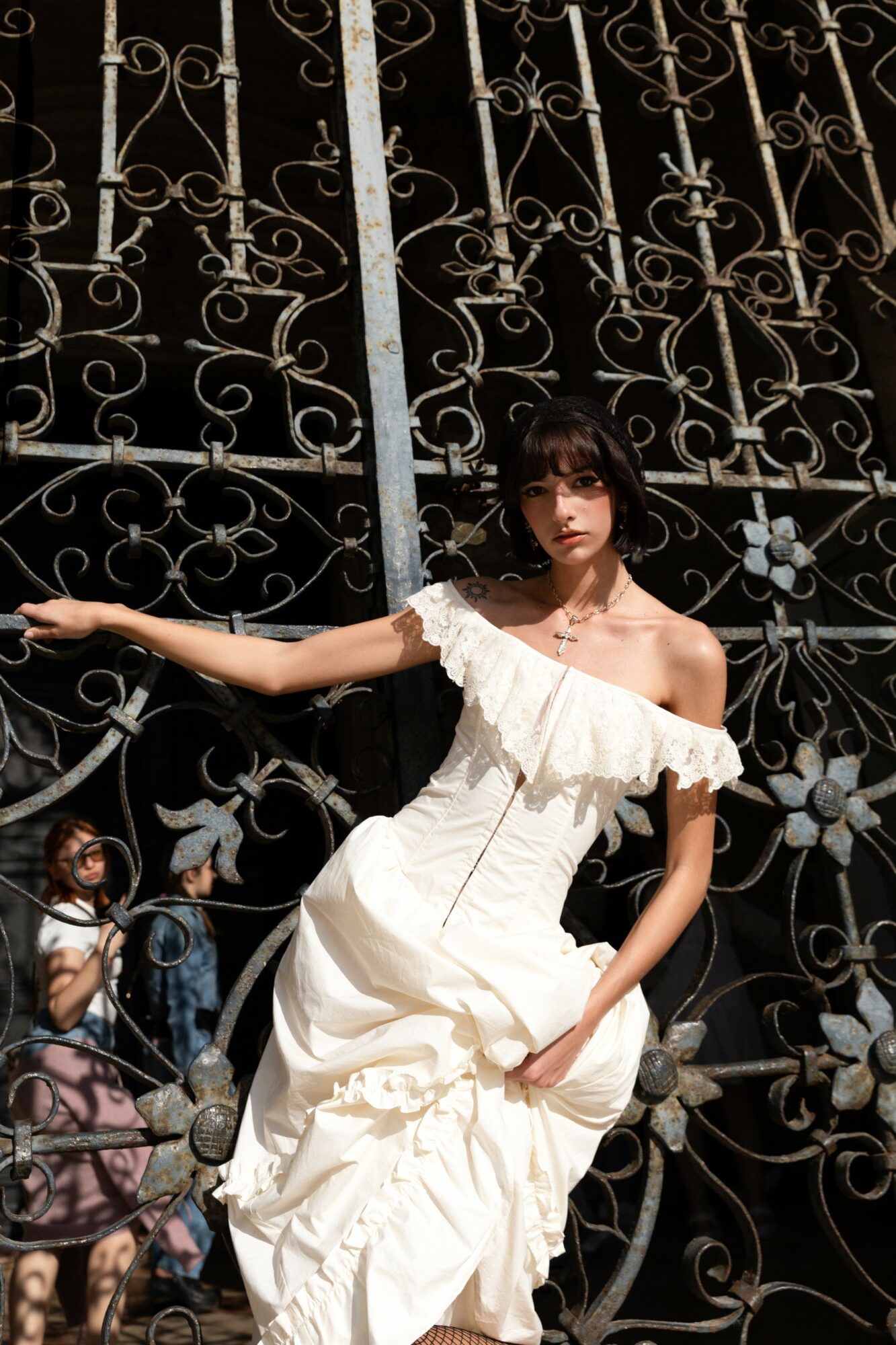 Young woman in a white dress leaning against a decorative metal gate, with children in the background.