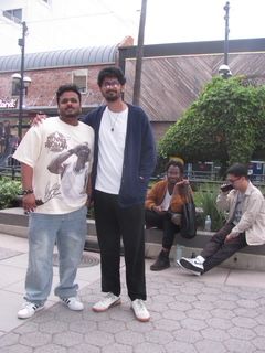 Two men and a woman standing outdoors near a bench and a bush, with buildings and a street in the background.