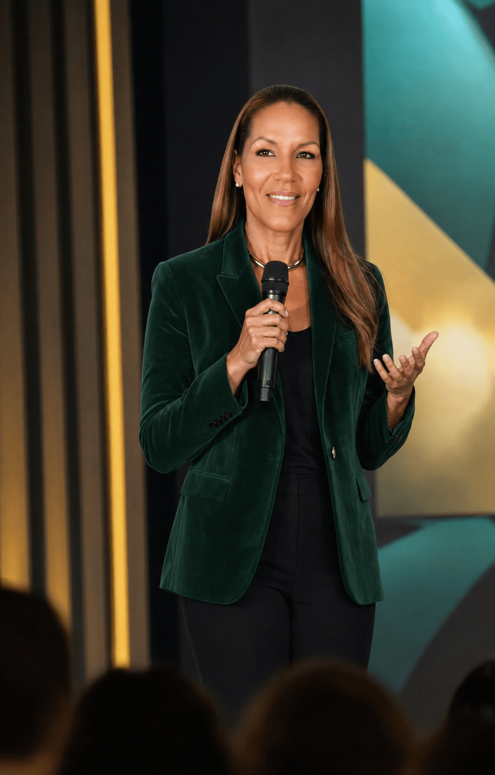 Woman speaking into a microphone on stage, wearing a dark green blazer and black outfit, with a colorful background.
