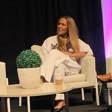 Woman with long hair sitting on a white armchair, holding a microphone, with a potted plant and a coffee cup on a table in front.