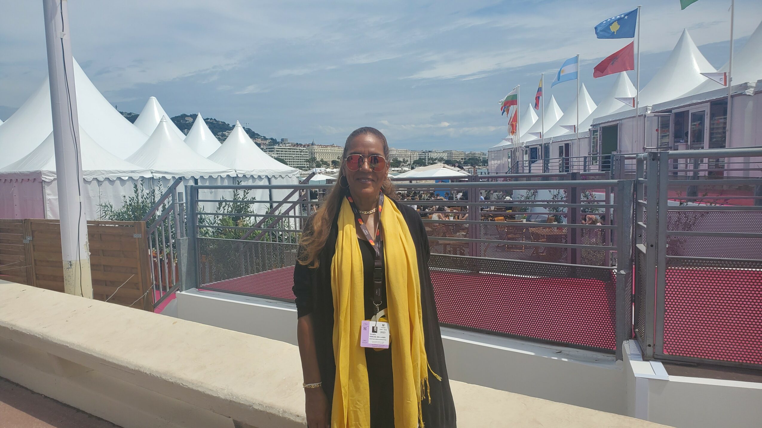 Person standing outdoors with tents and flags in the background, wearing a yellow scarf and black clothing.