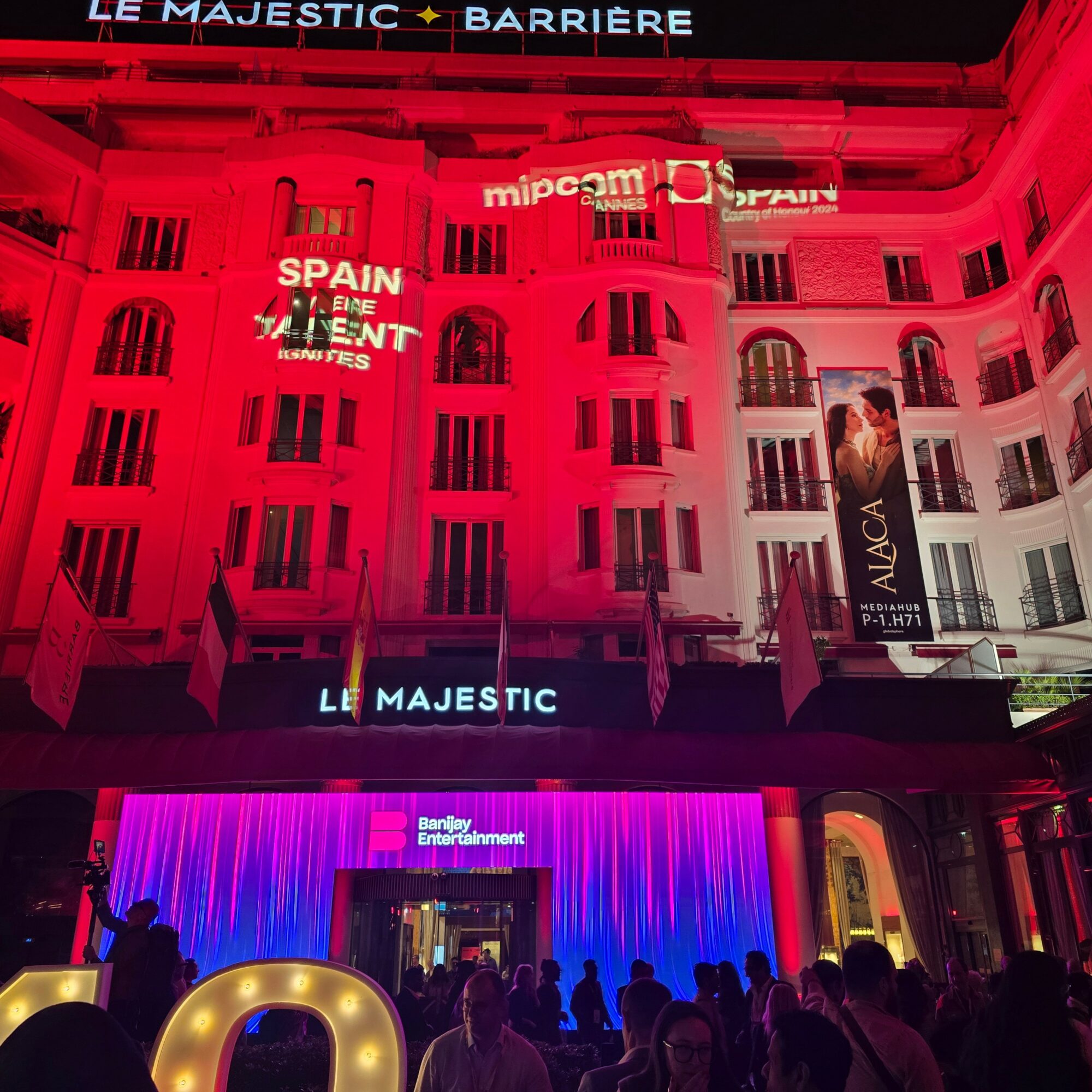 Night view of a building illuminated in pink and purple lights, with people gathered outside and signs on the building.