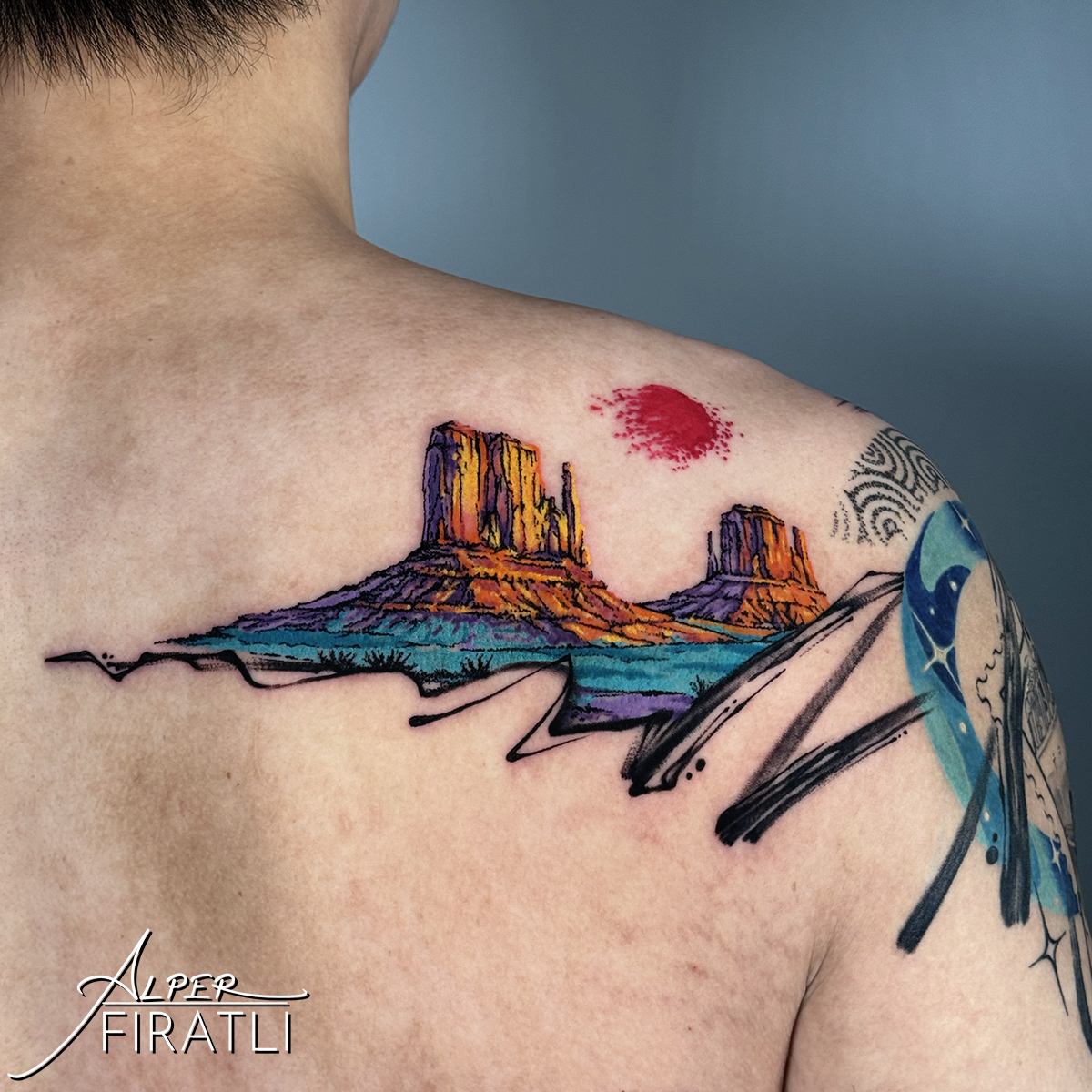 Colorful desert landscape tattoo on shoulder with red sun and abstract elements, signature in bottom left corner.