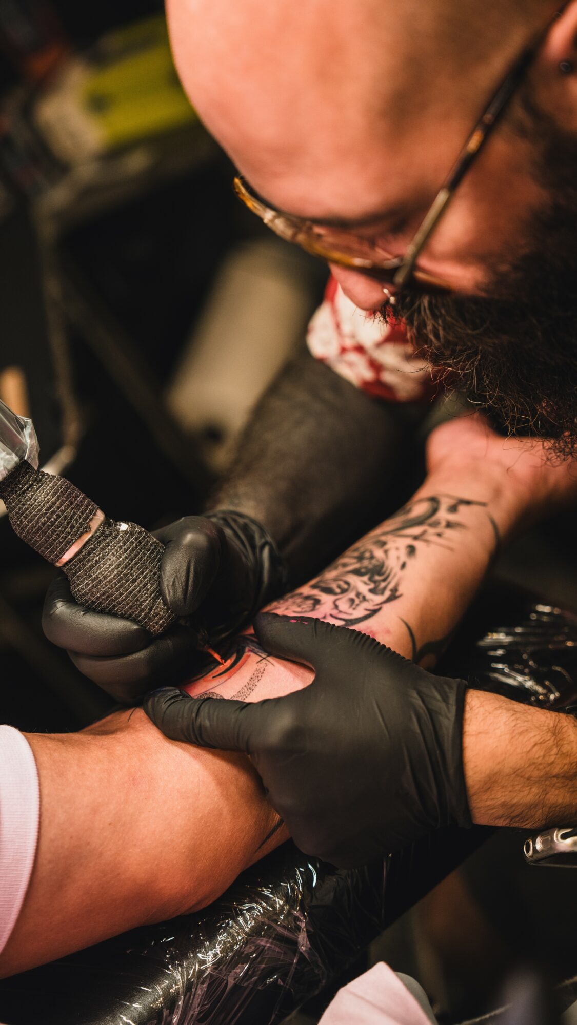 Tattooed arm being tattooed by a person wearing black gloves, with a tattoo machine in hand, in a tattoo studio.