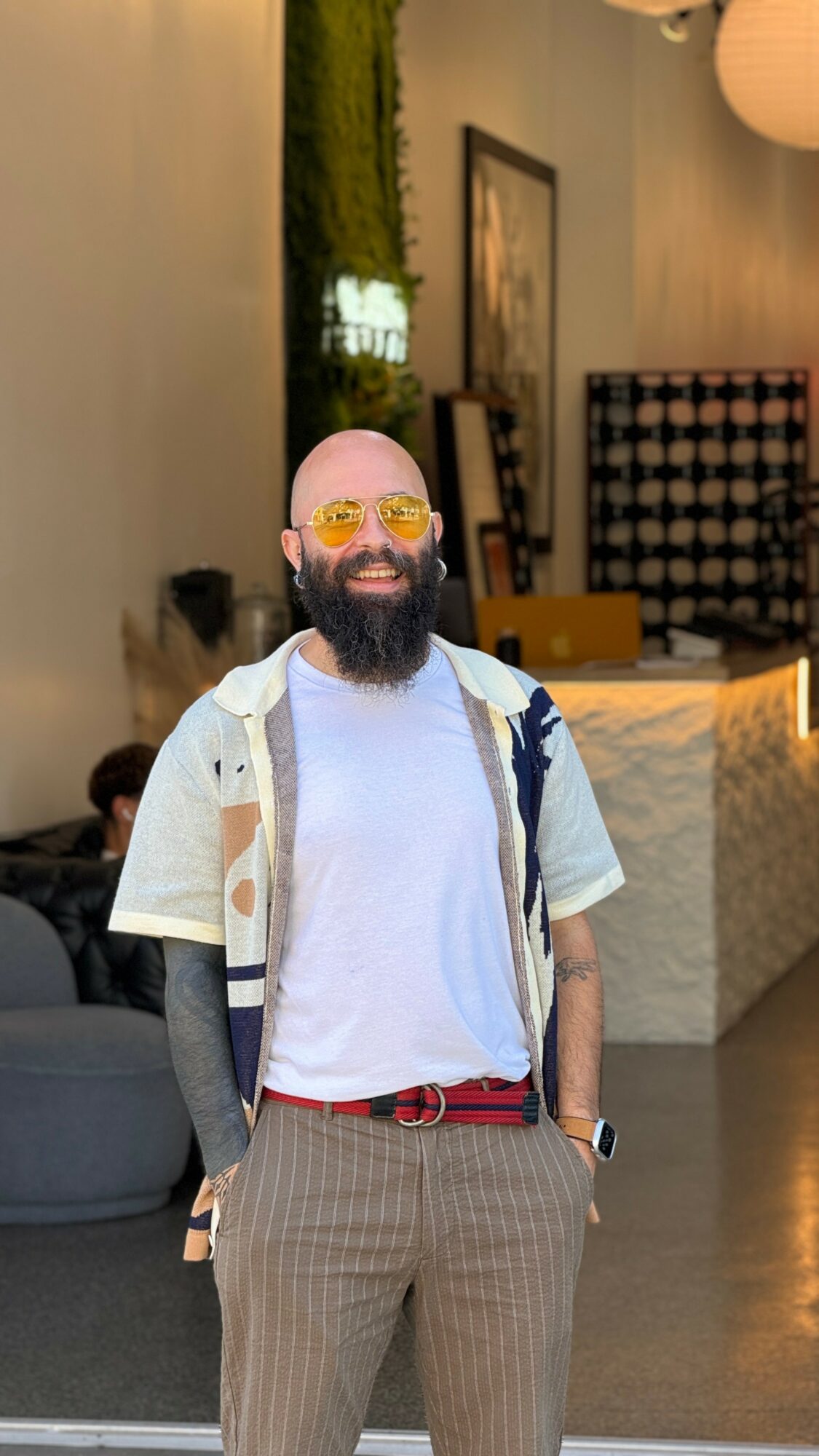 Bald man with a beard wearing glasses, a white t-shirt, and plaid pants, smiling indoors.