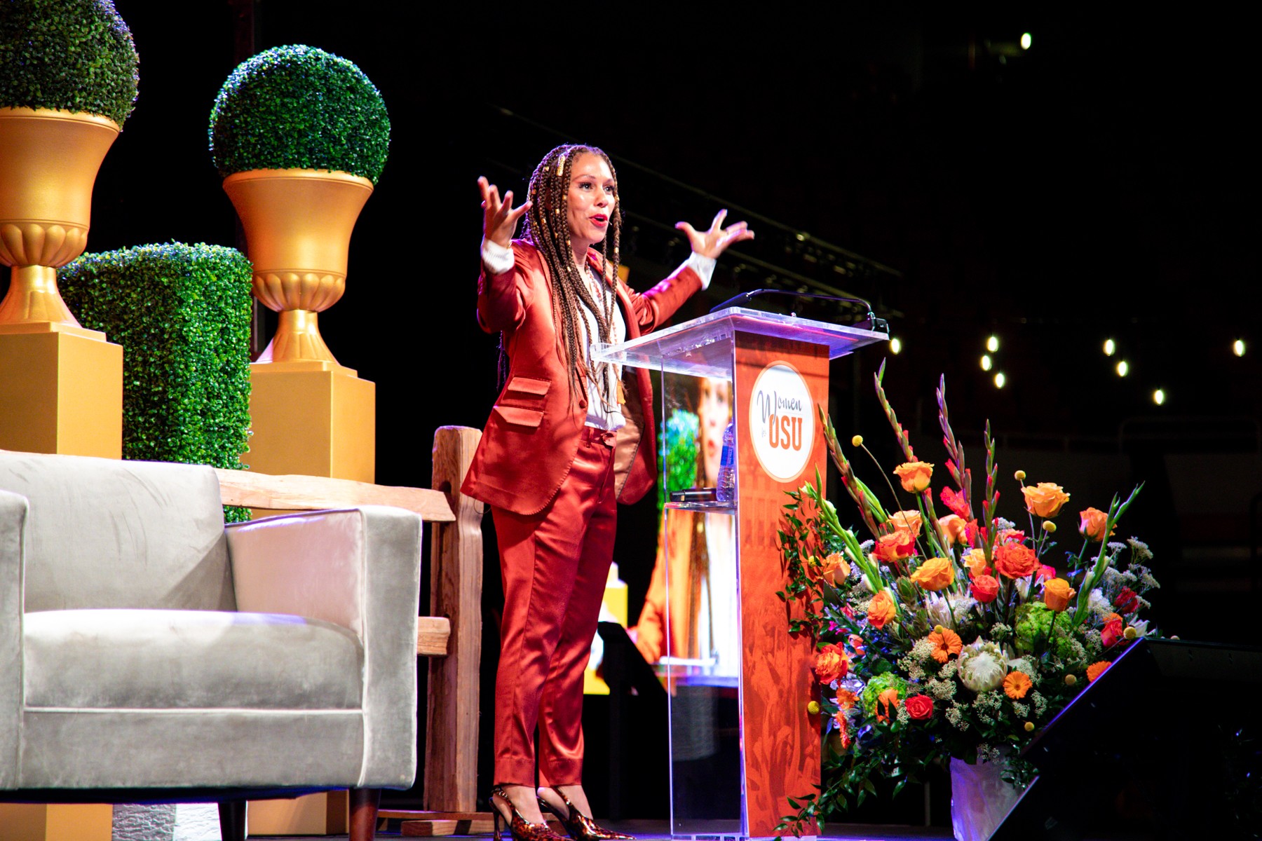 Young girl in red suit speaking at a podium with flowers and decorative plants behind her.