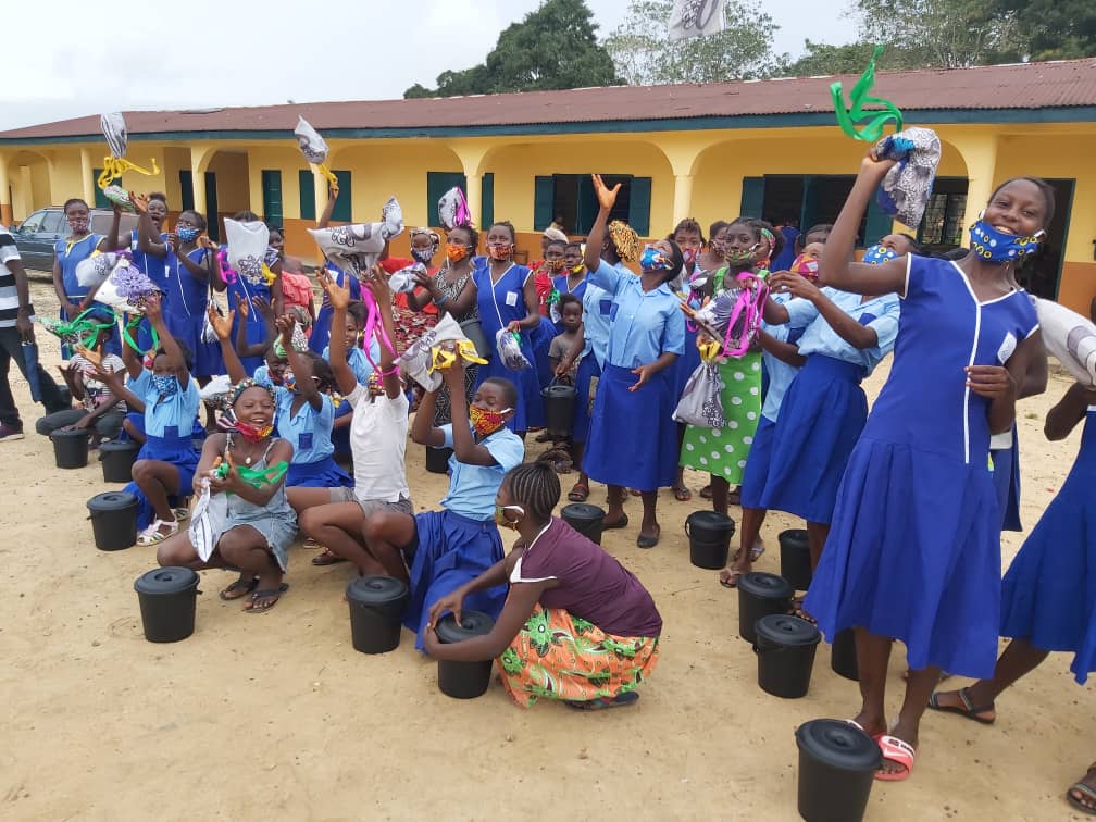 Group of children and women outdoors, some holding water bottles and buckets, in front of a yellow building, celebrating.