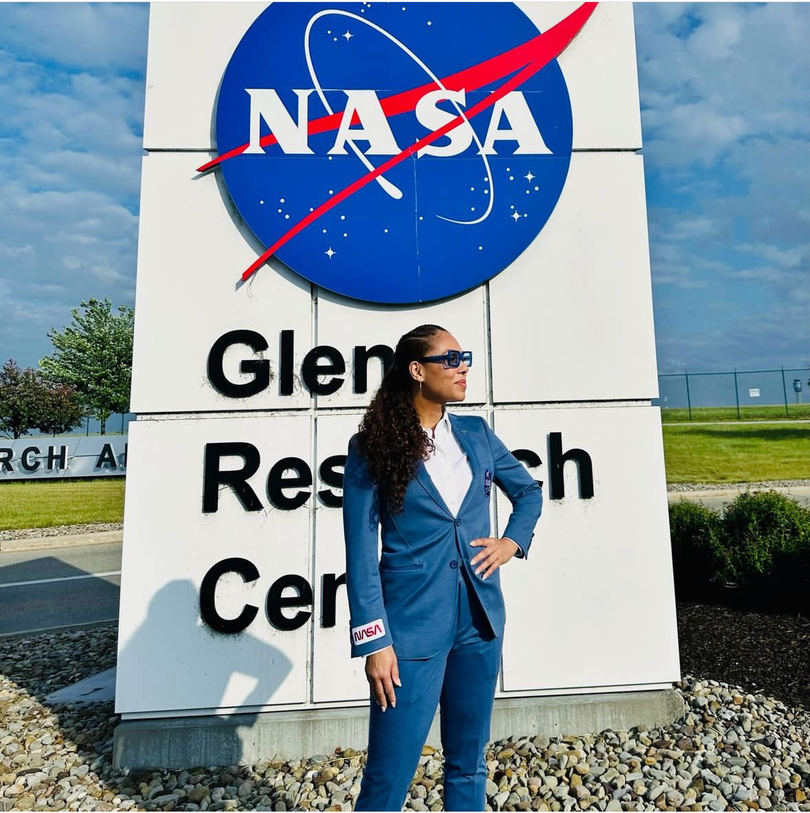 Woman in blue suit standing in front of NASA Glenn Research Center sign outdoors, with sky and greenery visible.