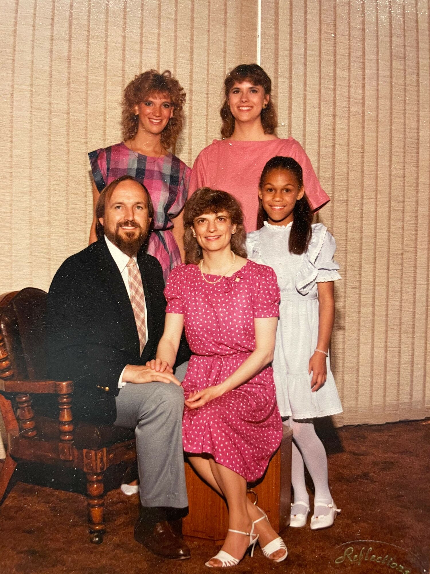 Group of five people, four women and one man, posing indoors in front of a beige curtain, smiling and dressed in casual and semi-formal clothing.