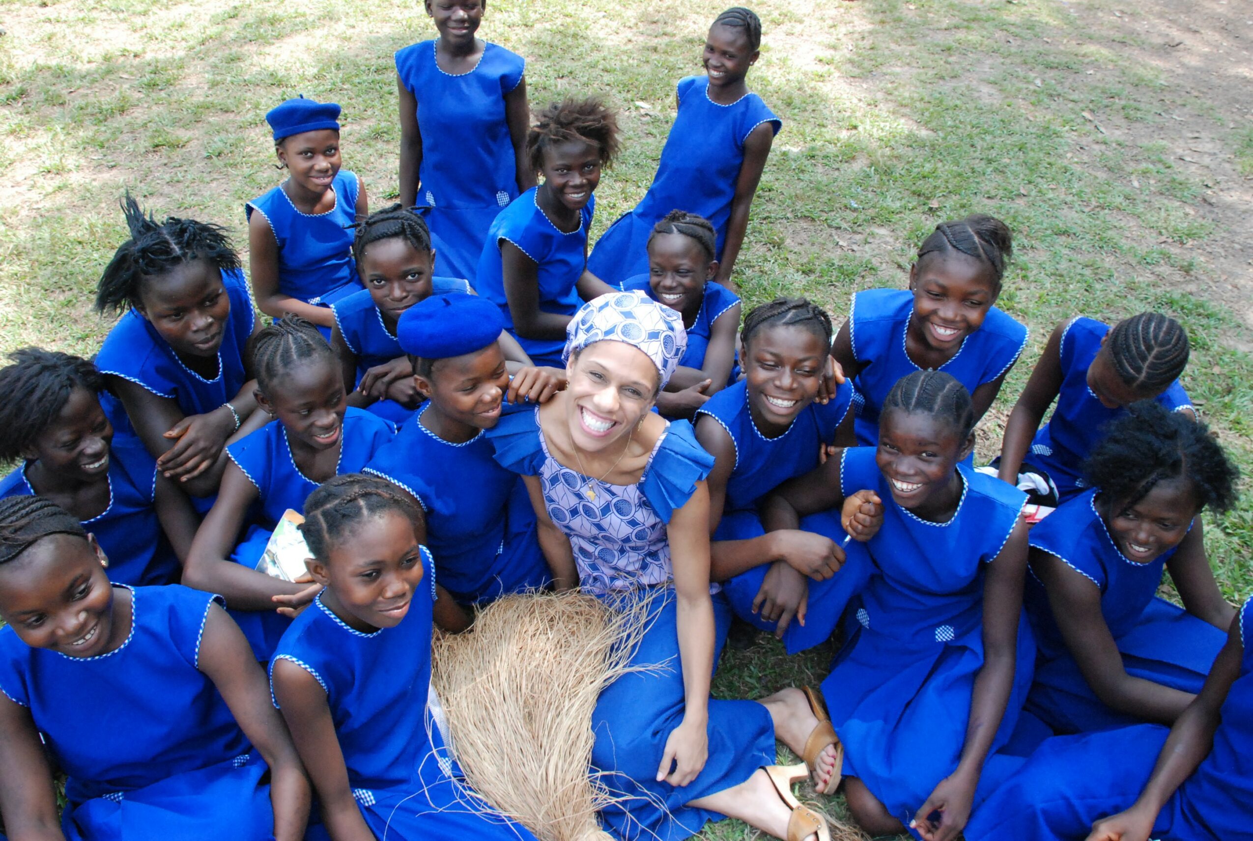 Group of children and a woman sitting outdoors on grass, all wearing blue clothing, smiling and posing for the photo.