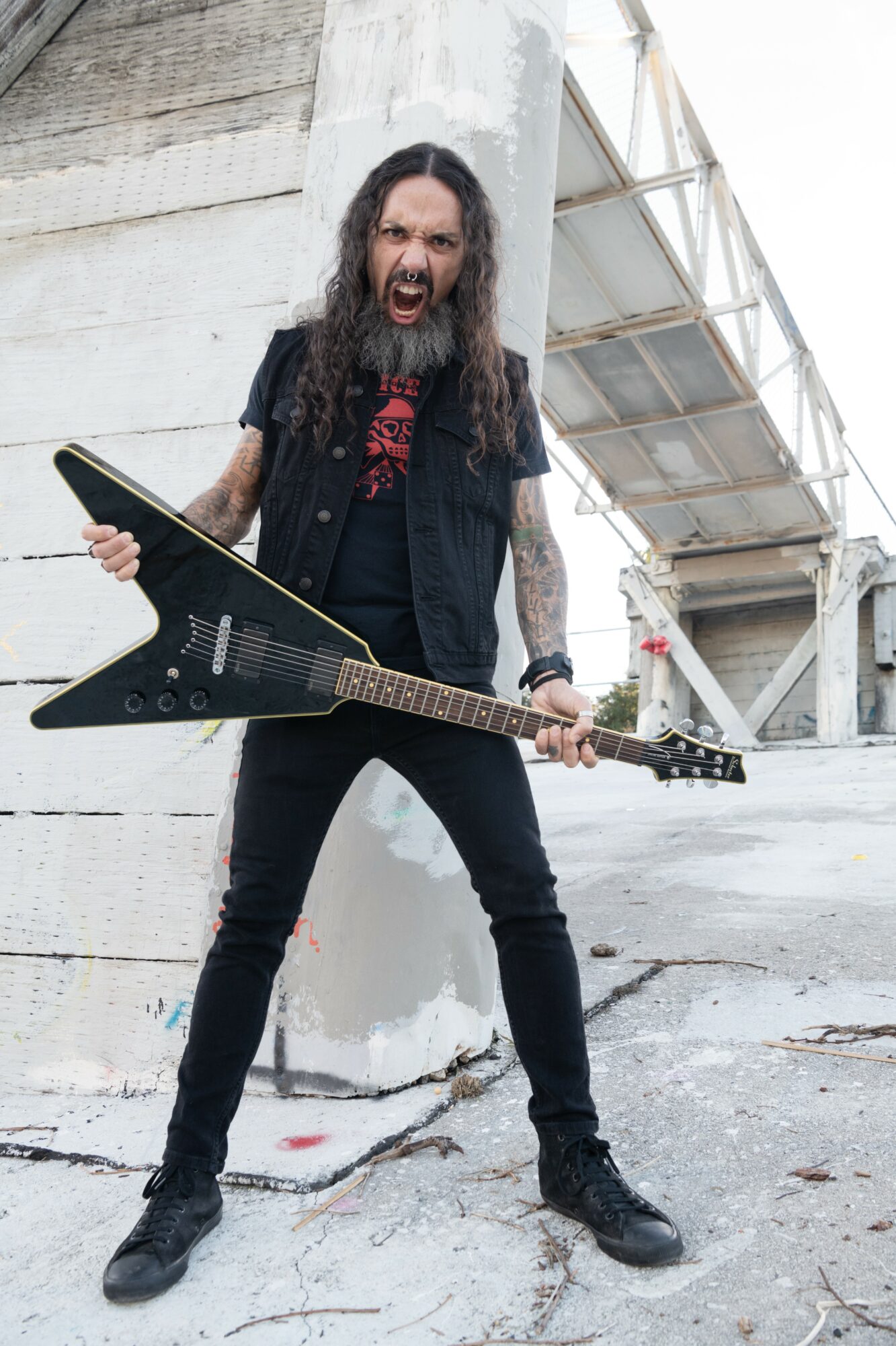 Man with long hair and beard holding an electric guitar, standing outdoors near a concrete structure.