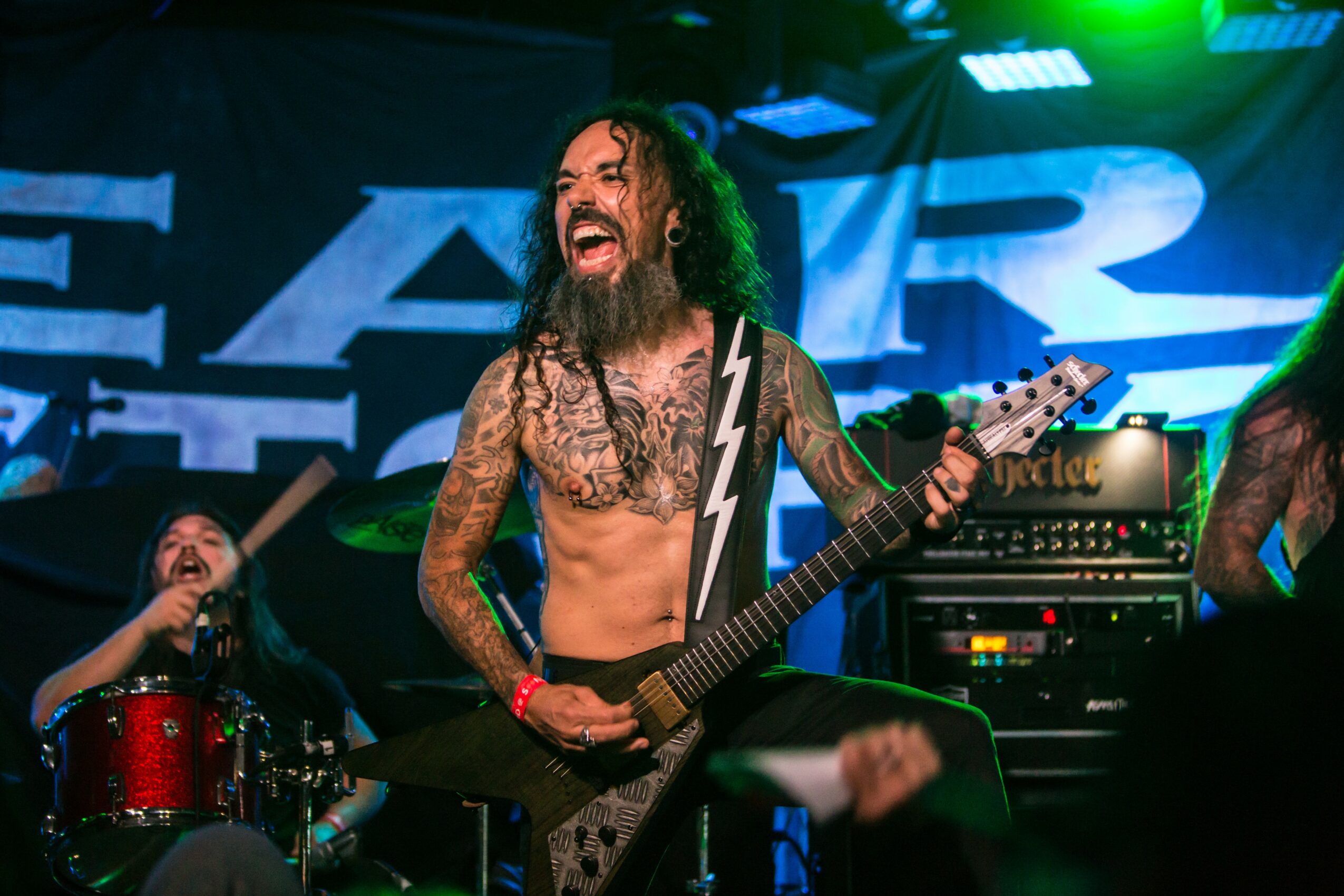 Musician with long curly hair and tattoos plays guitar on stage, smiling, with a drummer in the background, stage lights, and a large screen.