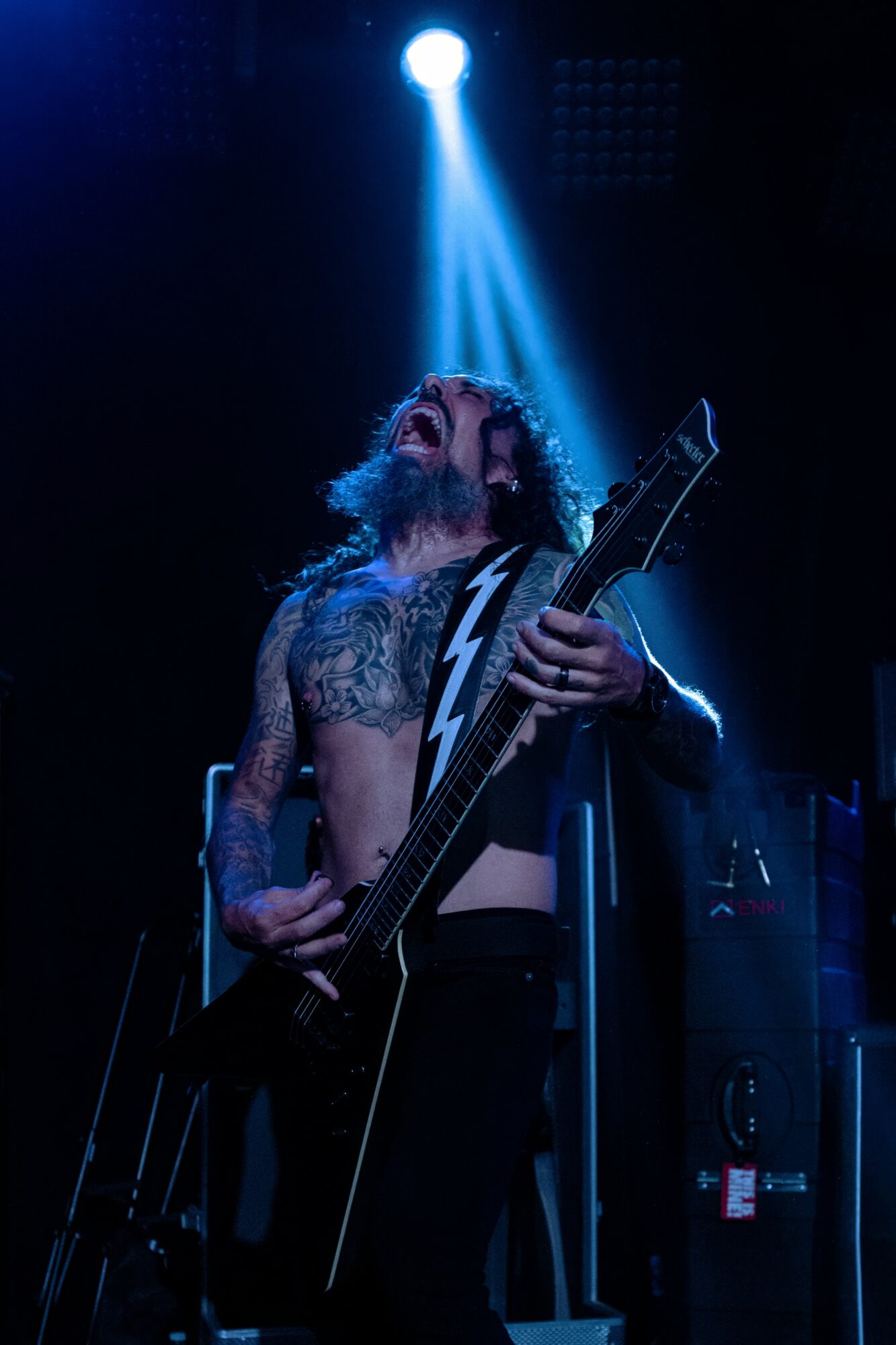 Musician with long hair and tattoos plays an electric guitar under a blue spotlight on stage.
