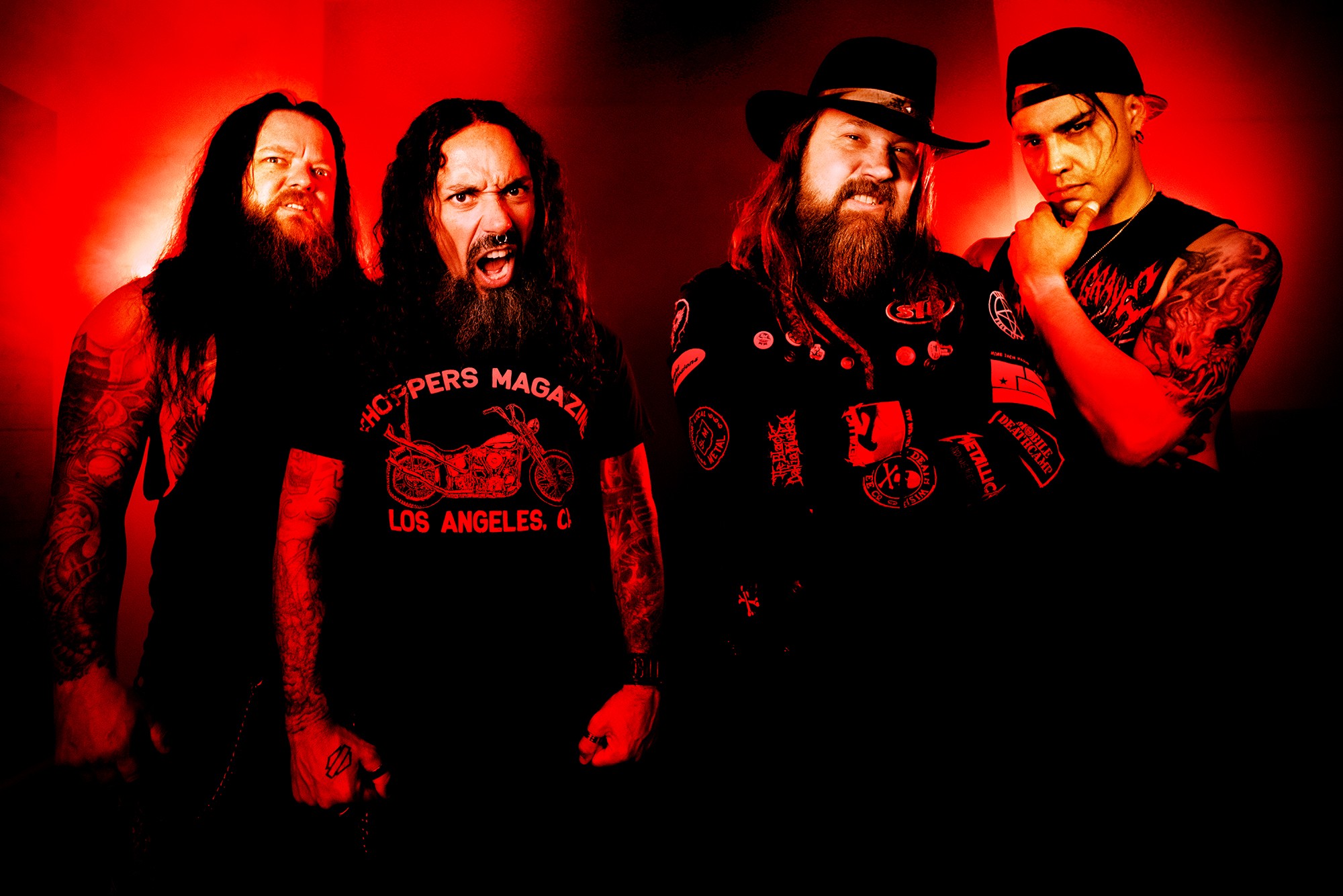 Four men with long hair and beards pose against a red background, some wearing hats and tattoos visible.