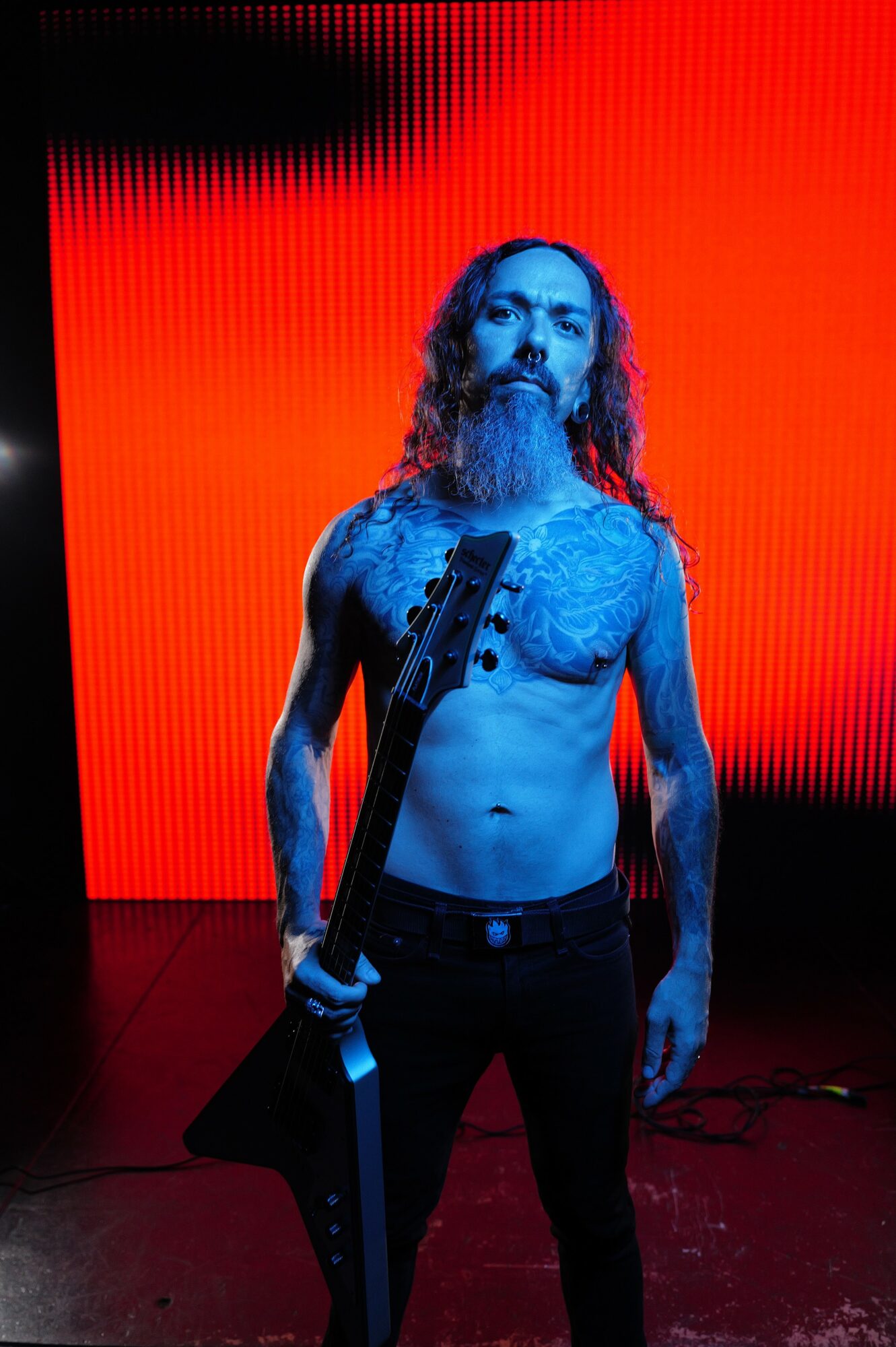 Man with long hair and beard holding a guitar, shirtless, standing against a red background.