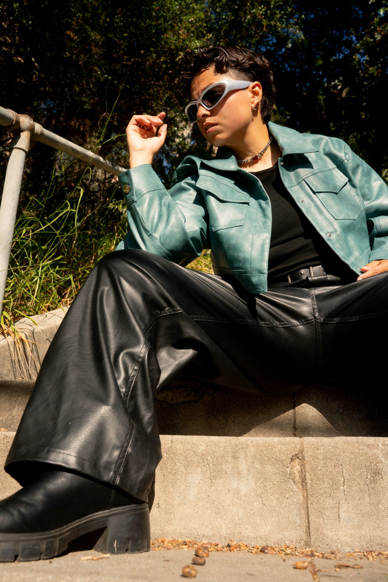 Person sitting outdoors on a concrete surface, wearing sunglasses, a green jacket, and black leather pants, with trees in background.