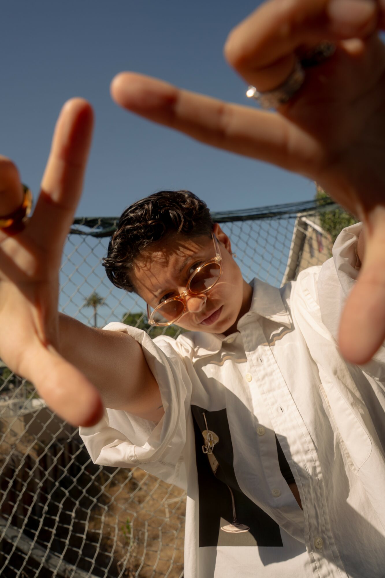 Young person with glasses and white shirt making a frame with hands outdoors, sunny day, fence in background.