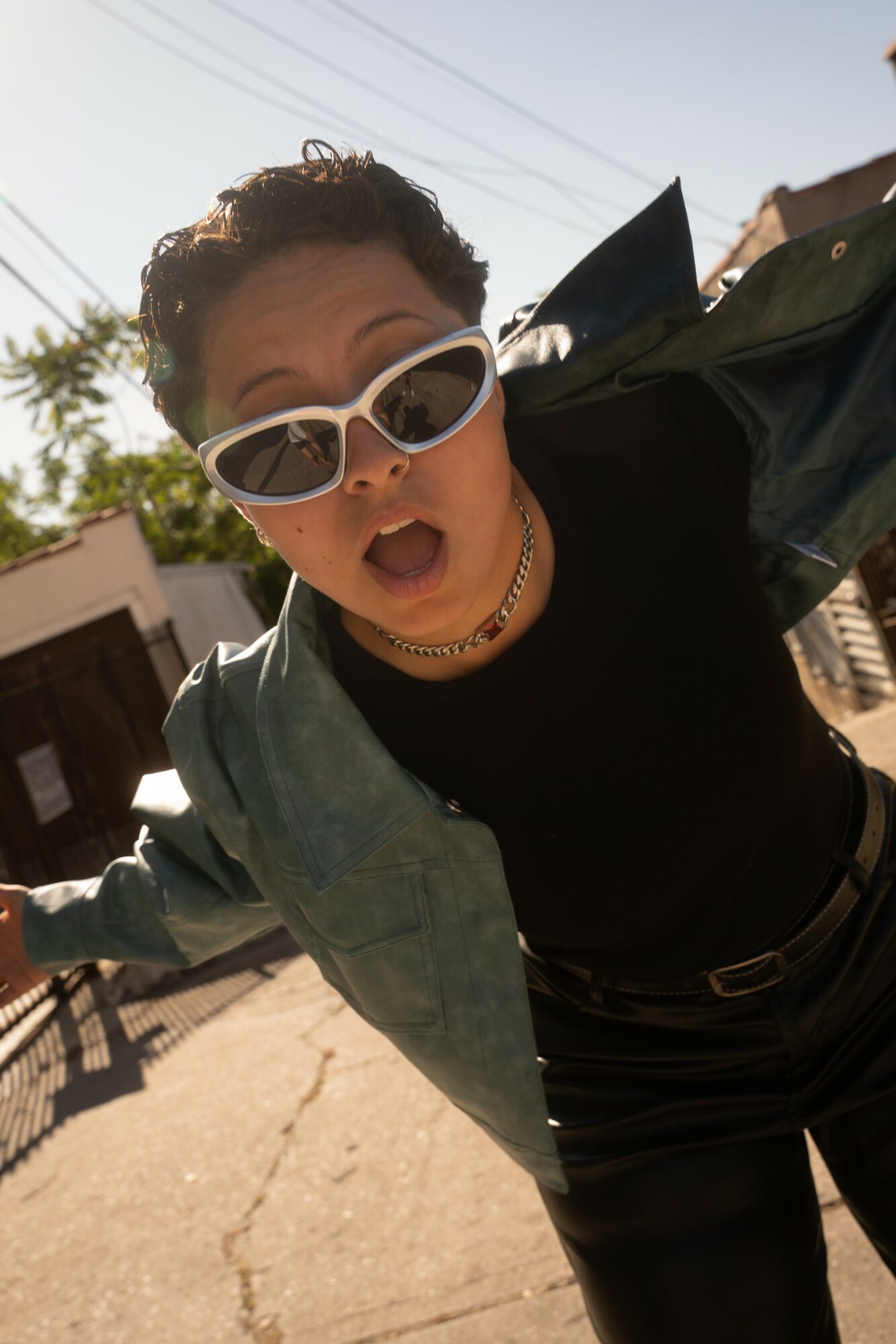 Person with short curly hair wearing sunglasses, black top, and green jacket outdoors, mouth open, leaning forward.