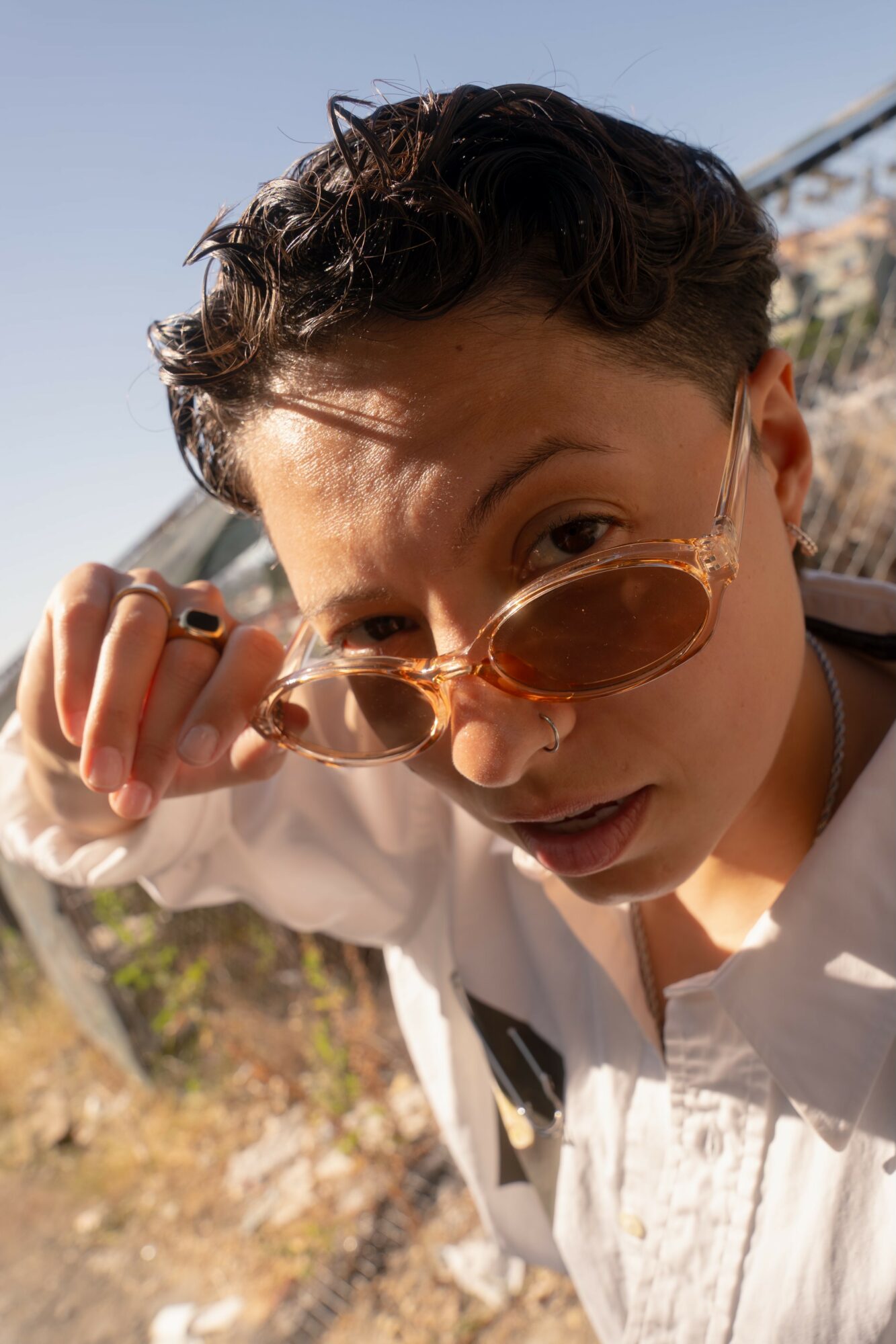 Person with short curly hair and sunglasses looking at camera outdoors, sunlight on face.