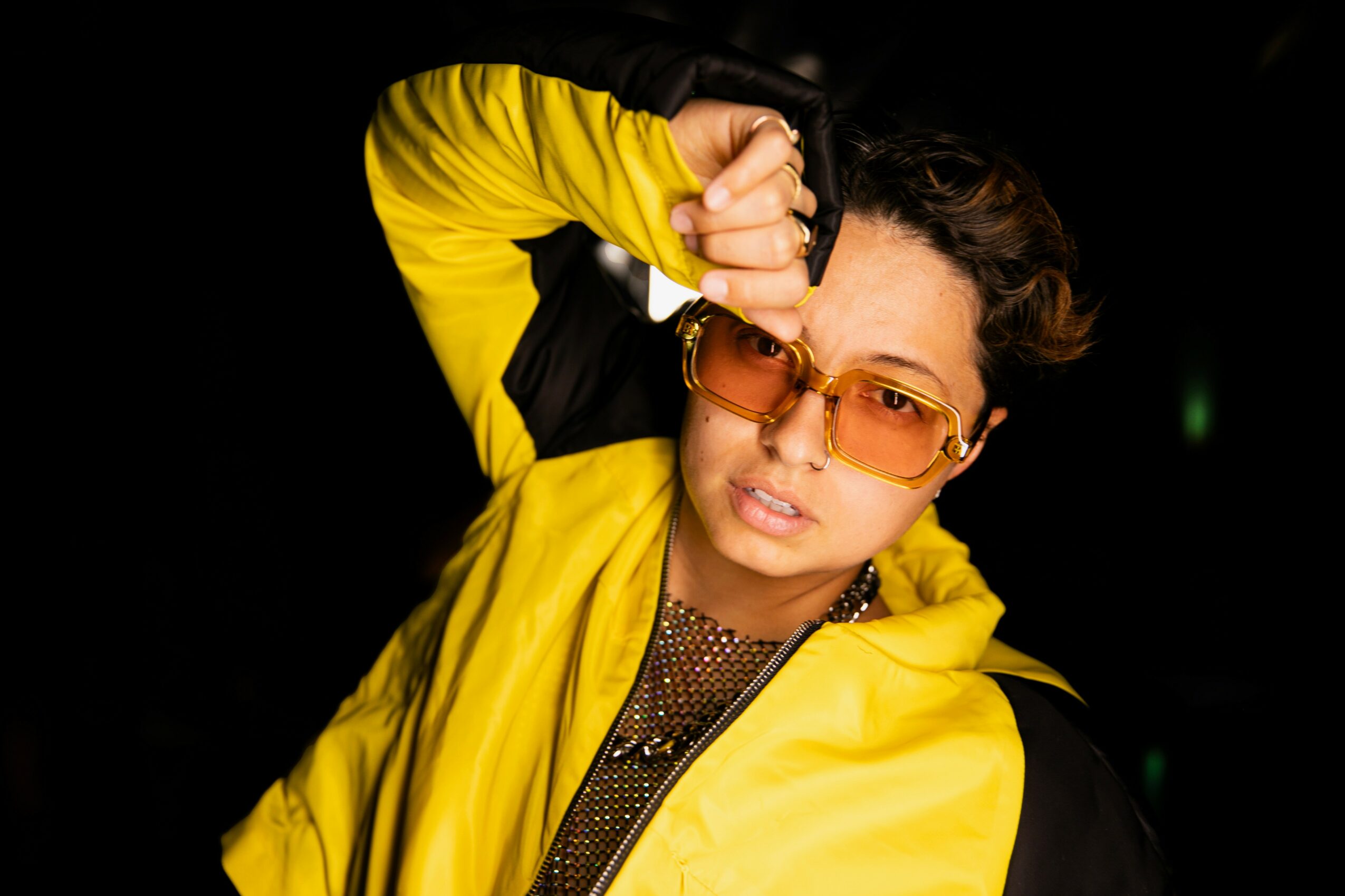 Person wearing yellow jacket and sunglasses, adjusting glasses with hand, against dark background.