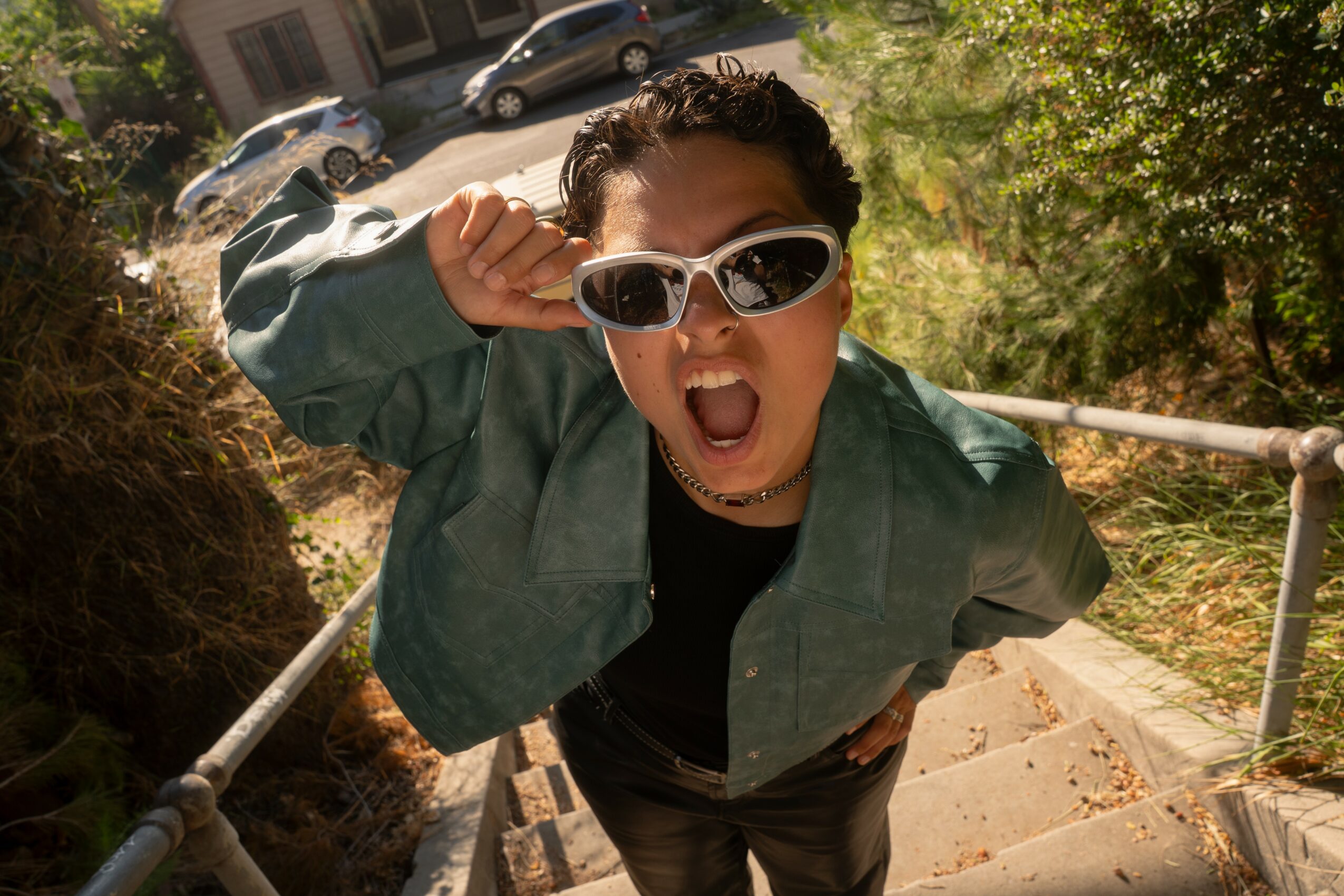 Person with sunglasses and green jacket shouting with mouth open, outdoors on stairs, trees and cars in background.