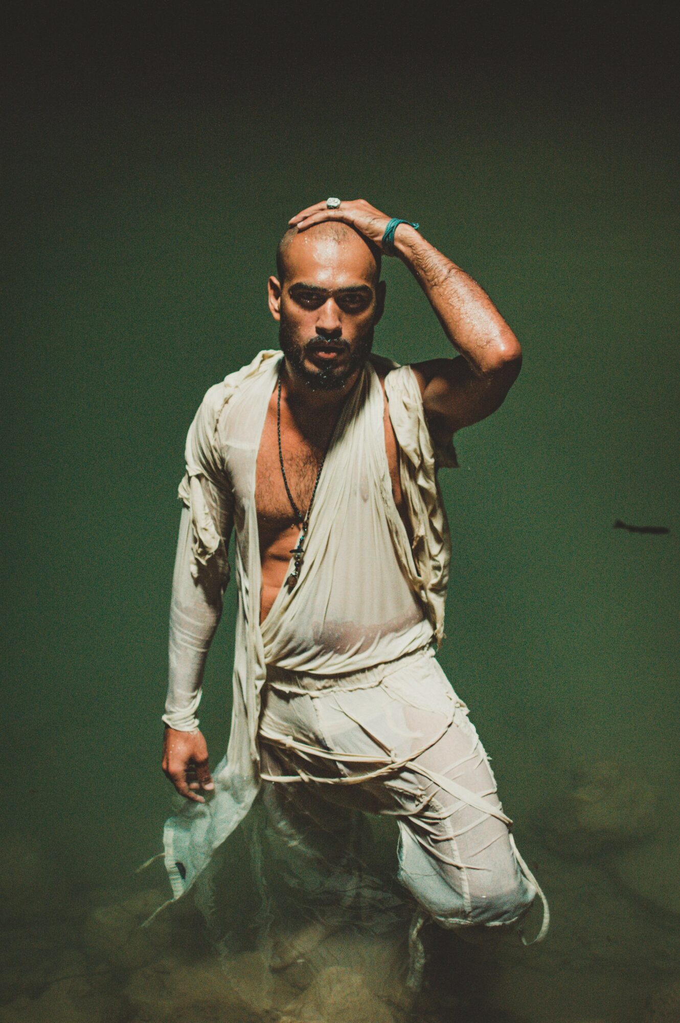 Man with a shaved head and beard, wearing a beige outfit, kneeling in water with a green background.