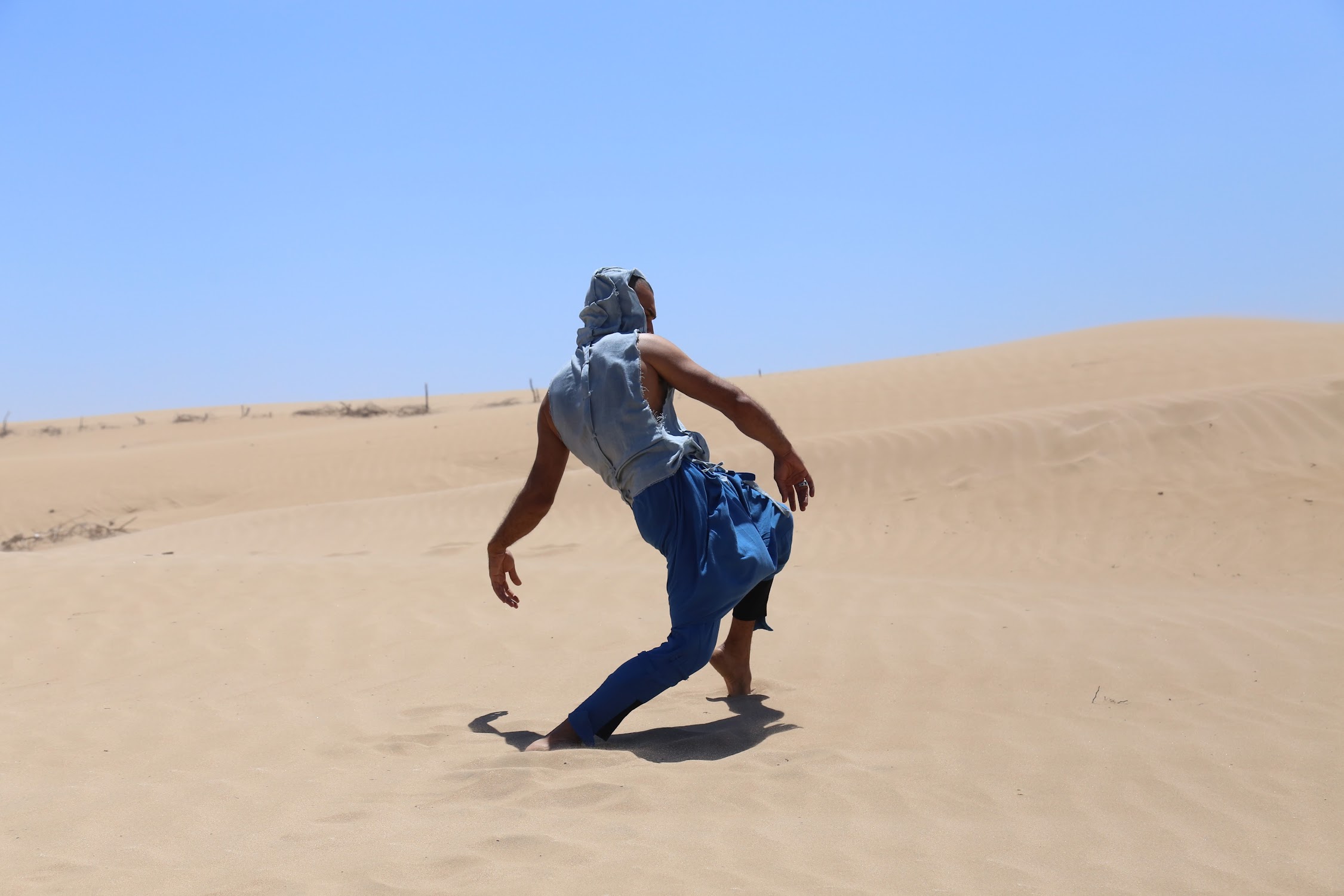 Person walking in a sandy desert landscape under a clear blue sky, wearing a head covering and casual clothing.
