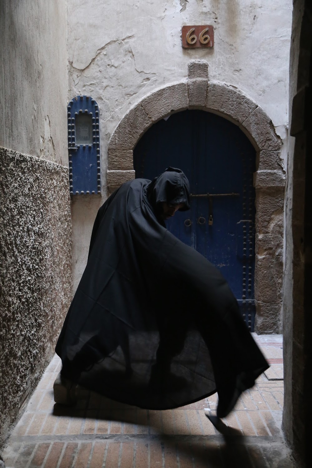 Person in black cloak bending forward in narrow alley with arched door and textured walls.