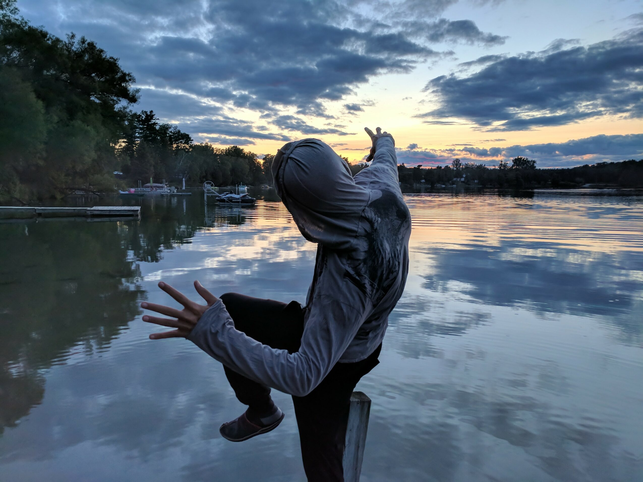 Person in hoodie sitting on a dock by a calm lake during sunset, with trees and clouds reflected in the water.