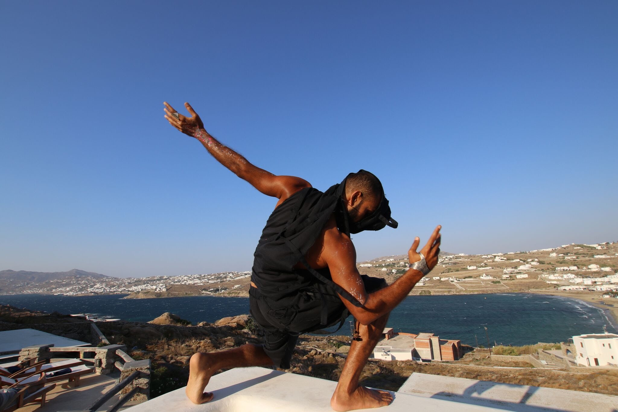 Person in black clothing balancing on one leg with arms extended, outdoors near water and buildings, clear sky.