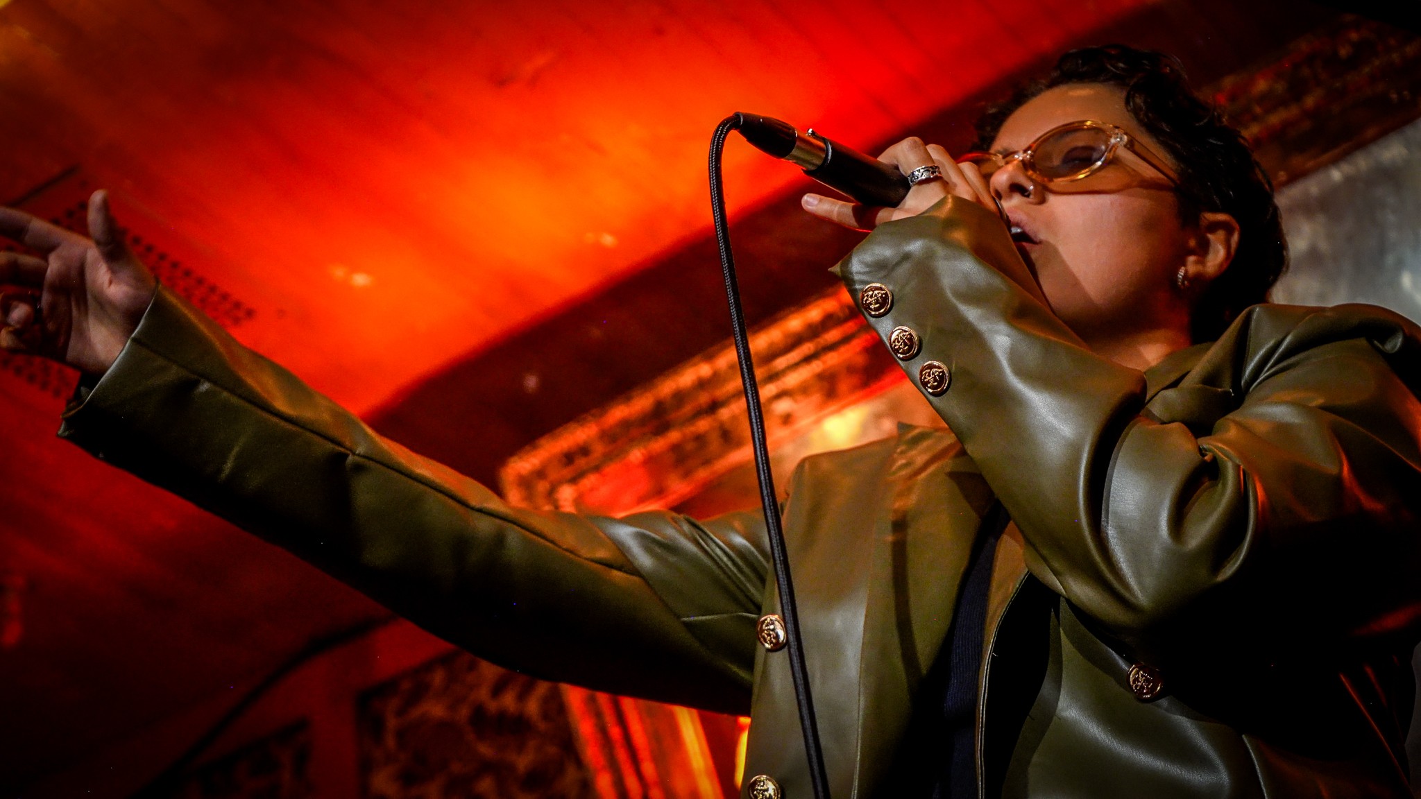 Performer singing into microphone with arm extended, stage lighting in background, wearing glasses and a jacket.