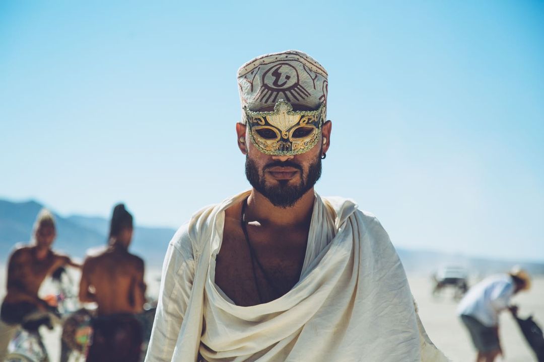 Man with beard wearing sunglasses, a patterned cap, and a white draped garment at the beach with people in the background.