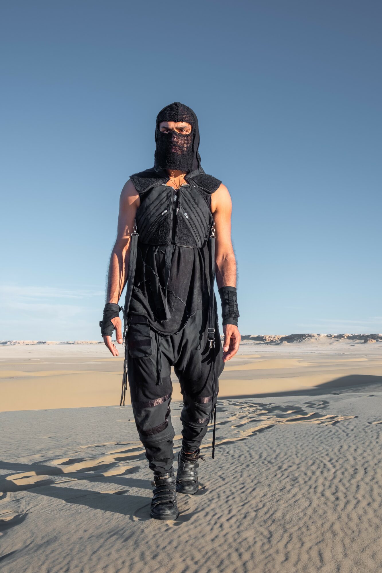 Person dressed in black walking on sandy beach with clear blue sky, wearing a face covering and gloves.