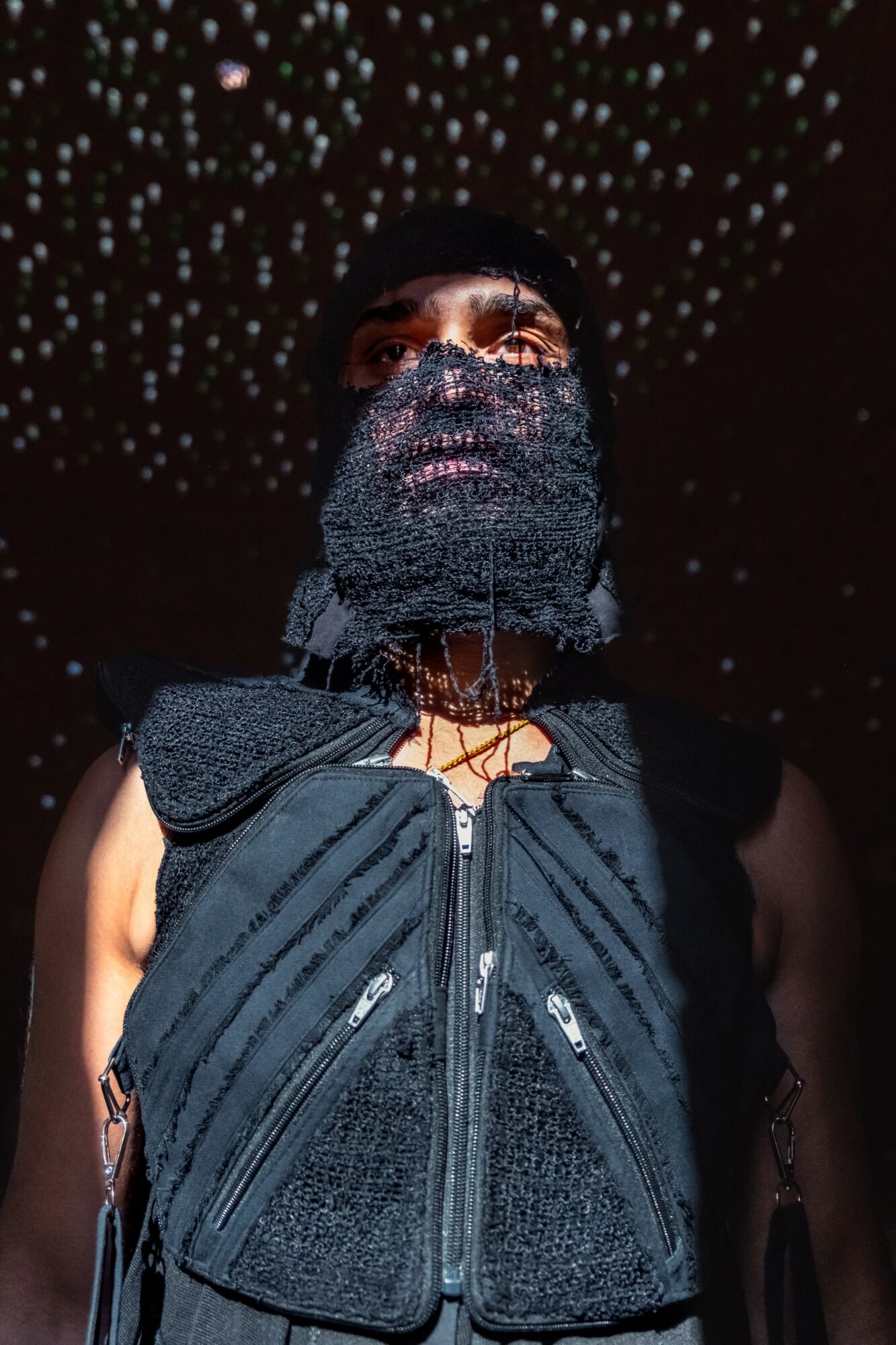 Person wearing a black face covering and vest, with a starry background overhead.