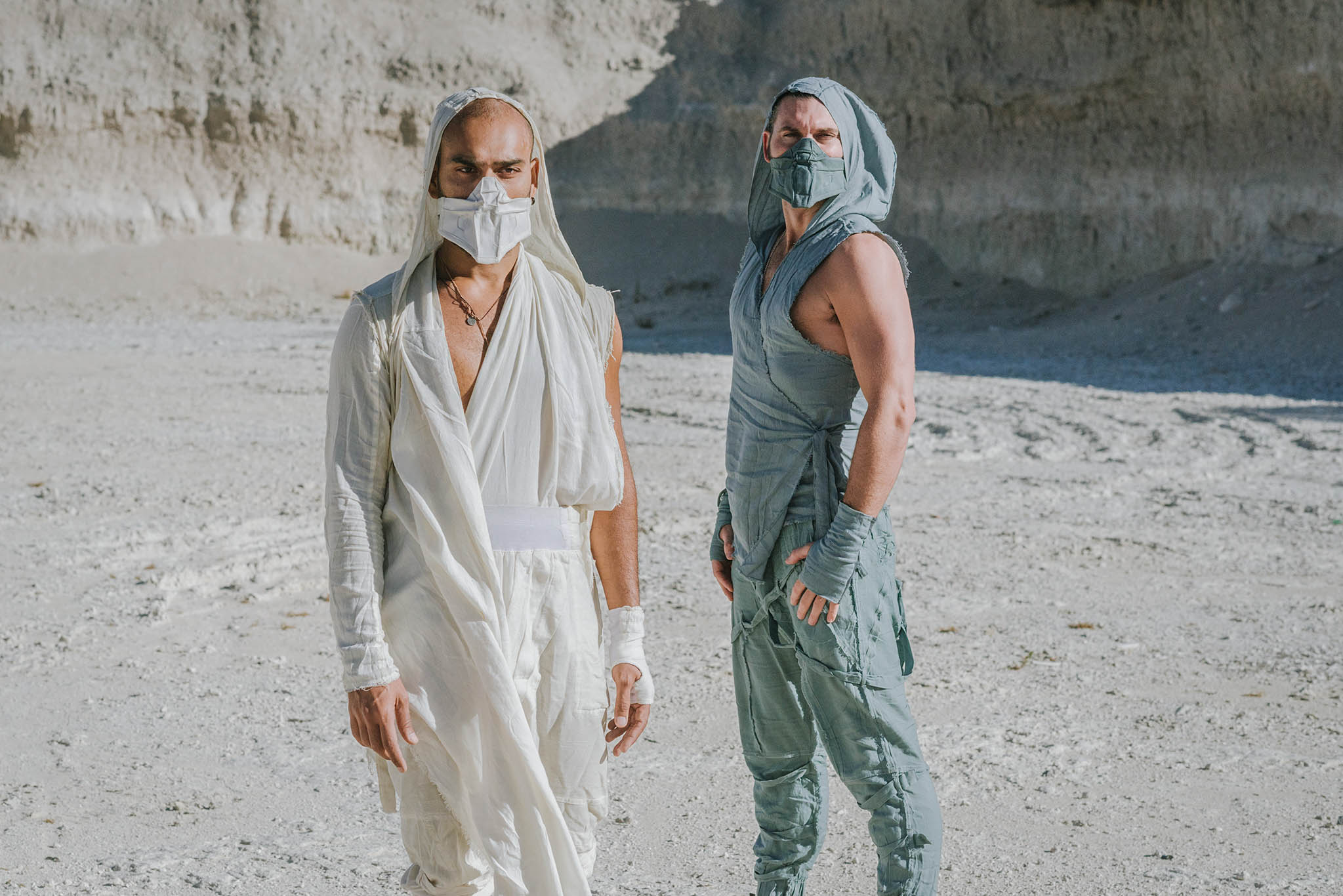 Two people wearing face masks and light-colored clothing stand outdoors on a sandy surface with a rocky background.