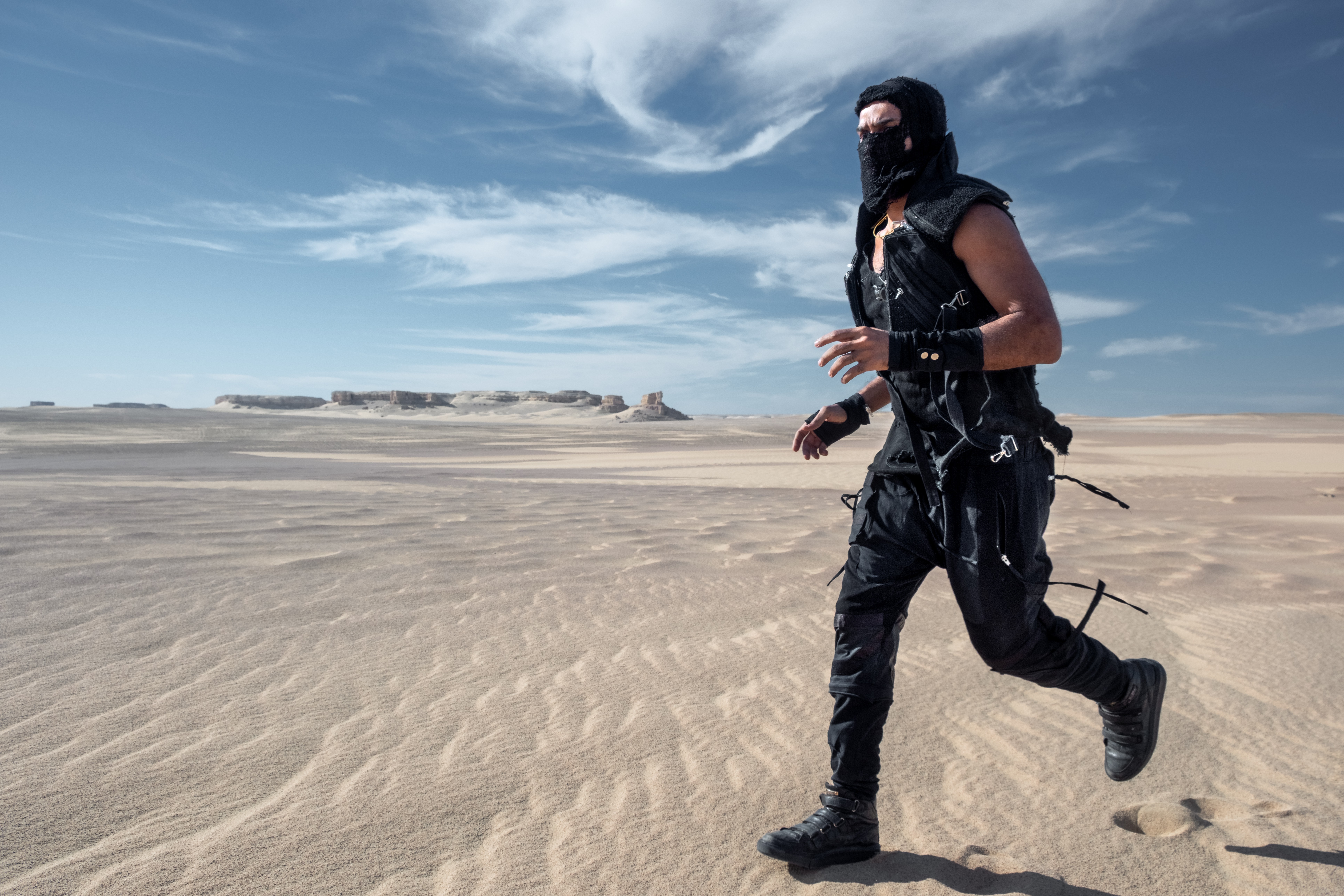 Person running on sandy beach with ocean and sky in background, wearing black tactical gear and mask.