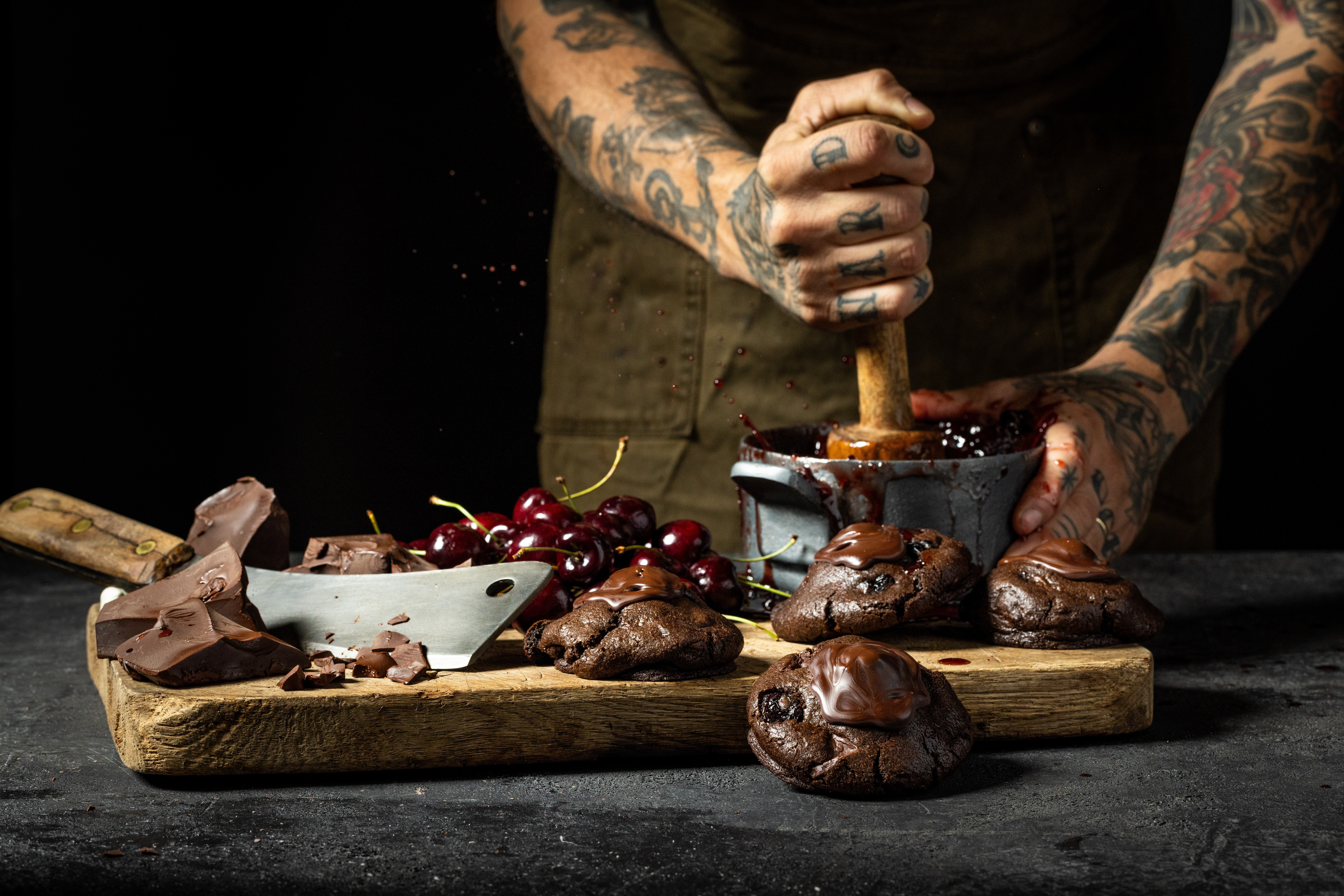 Person with tattooed arms mashing cooked food in a bowl on a wooden board, with cherries and chocolate nearby.