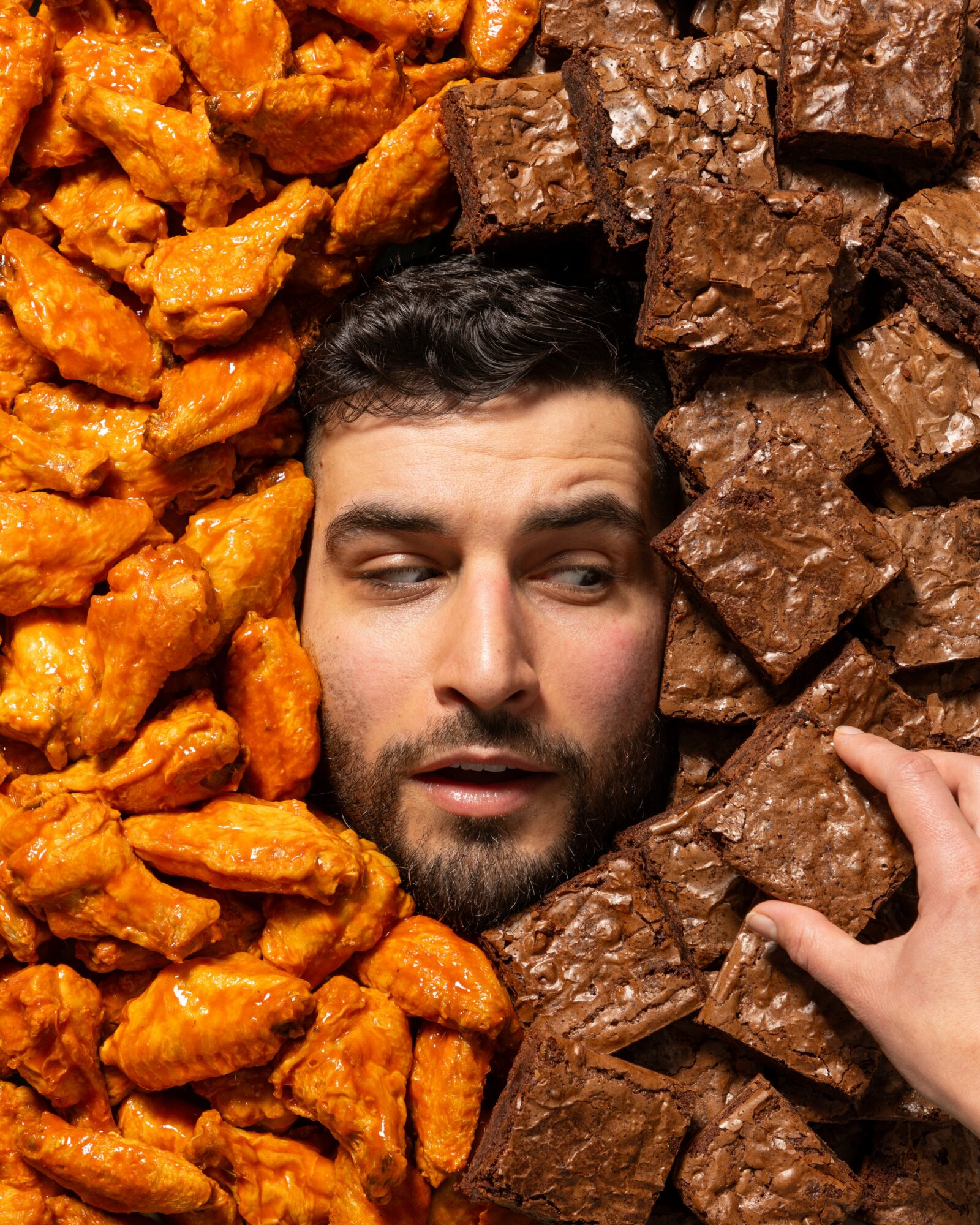 Person's face surrounded by orange sweet potatoes and brown chocolate chunks, with hand touching chocolate pieces.