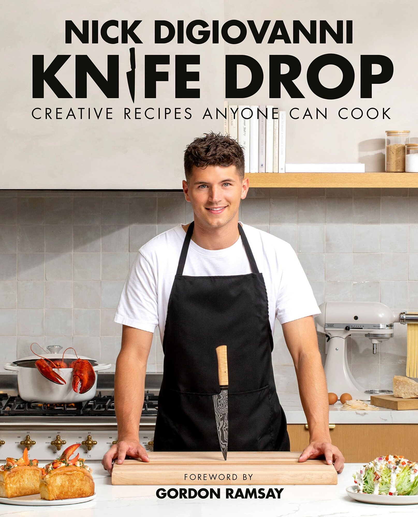 Young man in a kitchen wearing a black apron, smiling, with baked goods and kitchen utensils around him.