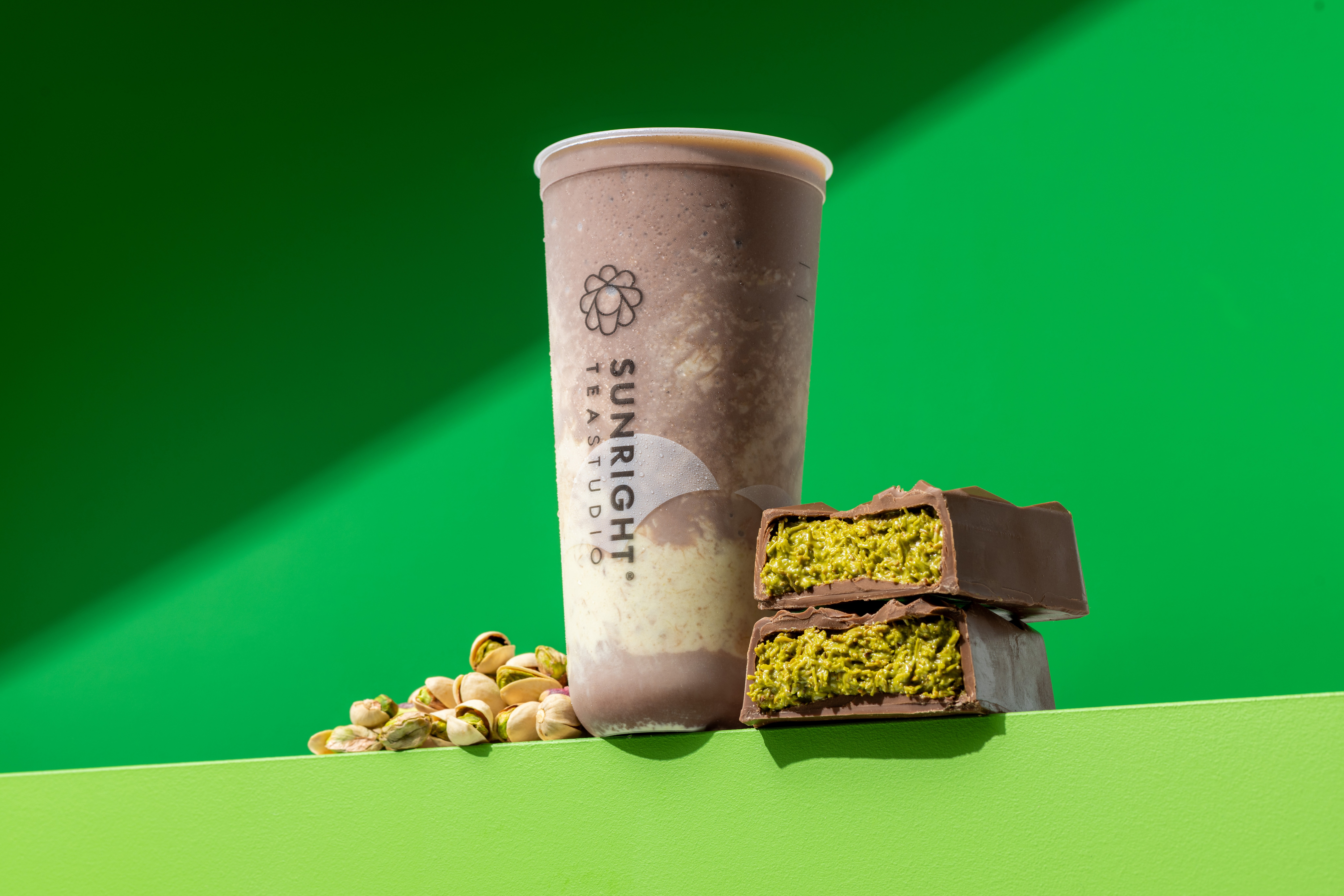 A beverage cup with a logo, two green layered cake slices with chocolate, and pistachios on a green background.