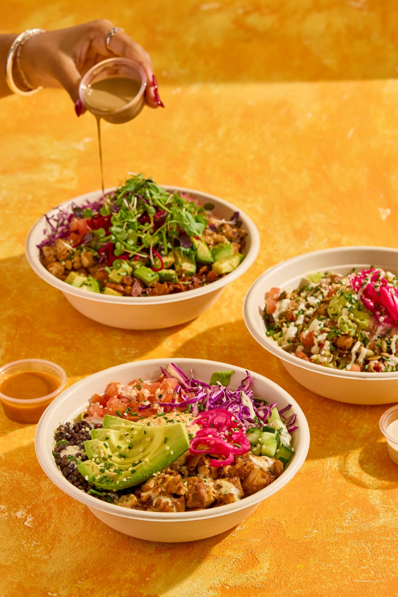 Hand pouring dressing over a bowl of salad with greens, avocado, and vegetables, on a yellow surface.