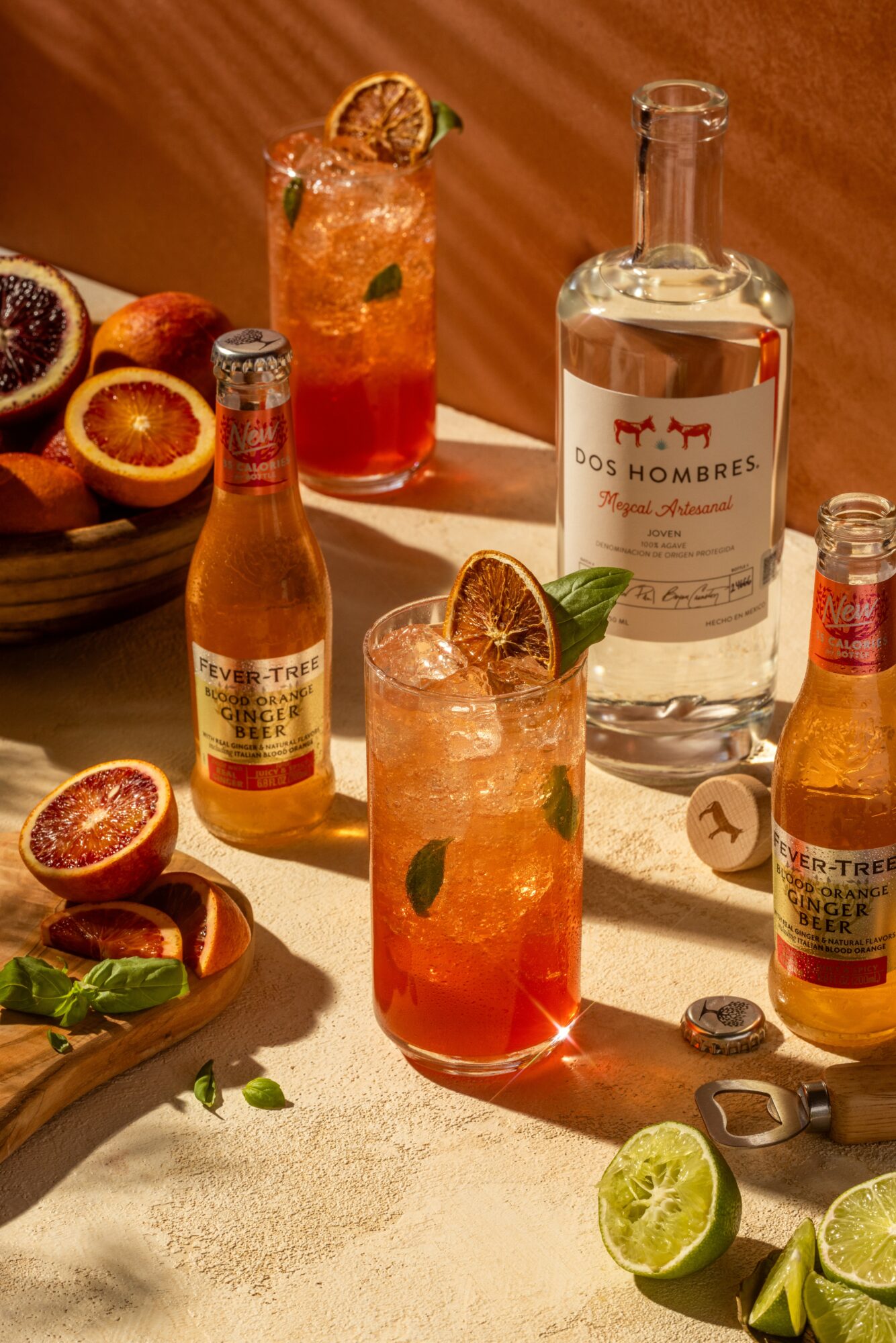 Two glasses of cocktail with ice, lime, and mint, surrounded by passion fruits, lime wedges, and bottles on a table.
