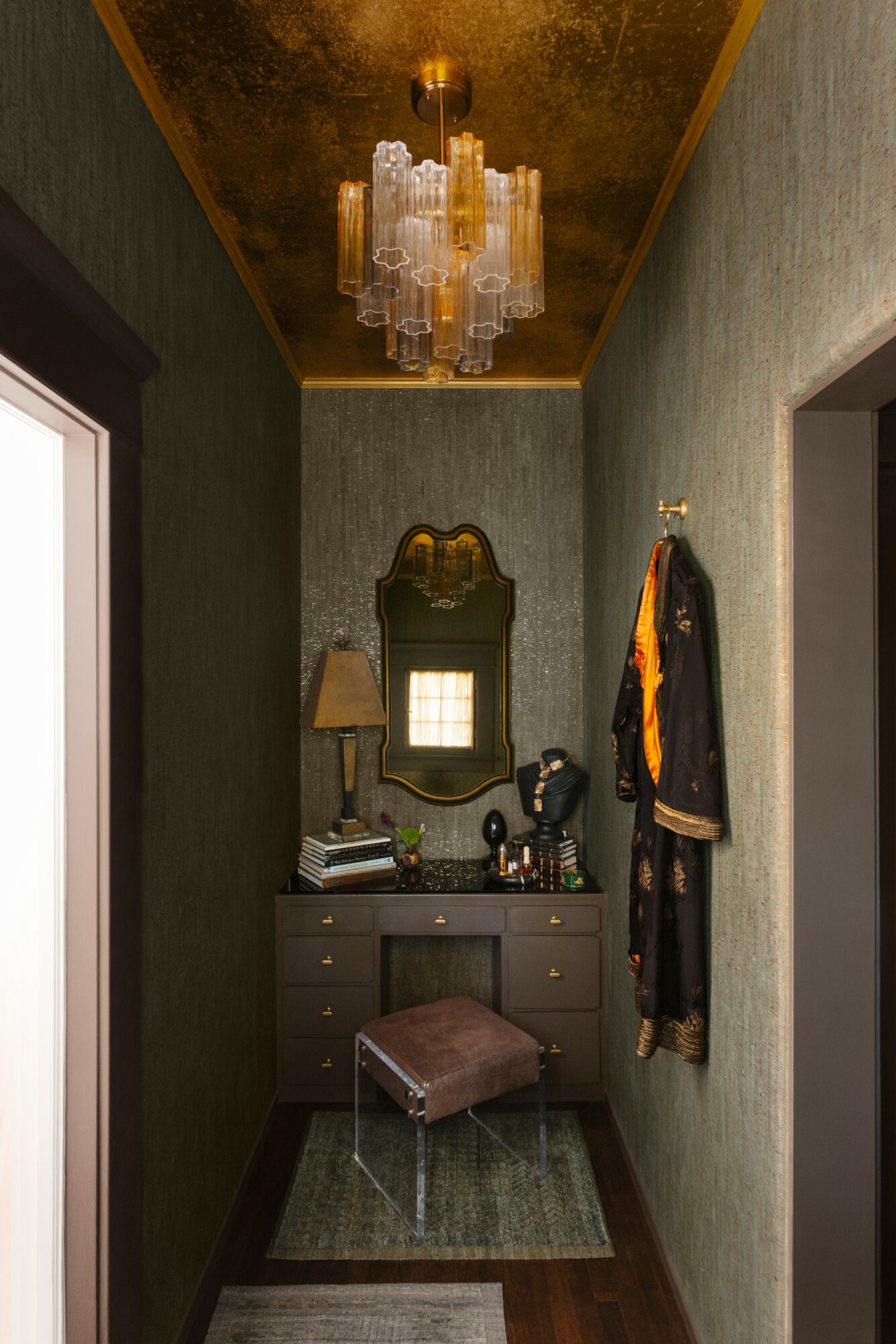 Small vanity area with mirror, lamp, and accessories, a stool, and a chandelier overhead, in a narrow hallway.