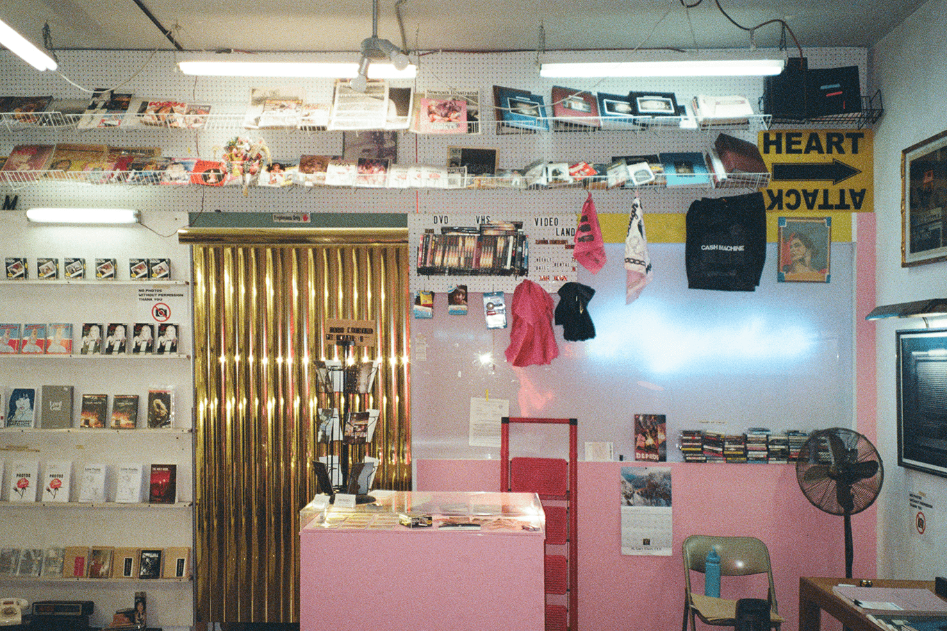 Interior of a store with shelves, hanging clothes, and a pink counter, illuminated by ceiling lights, with a television on the right.