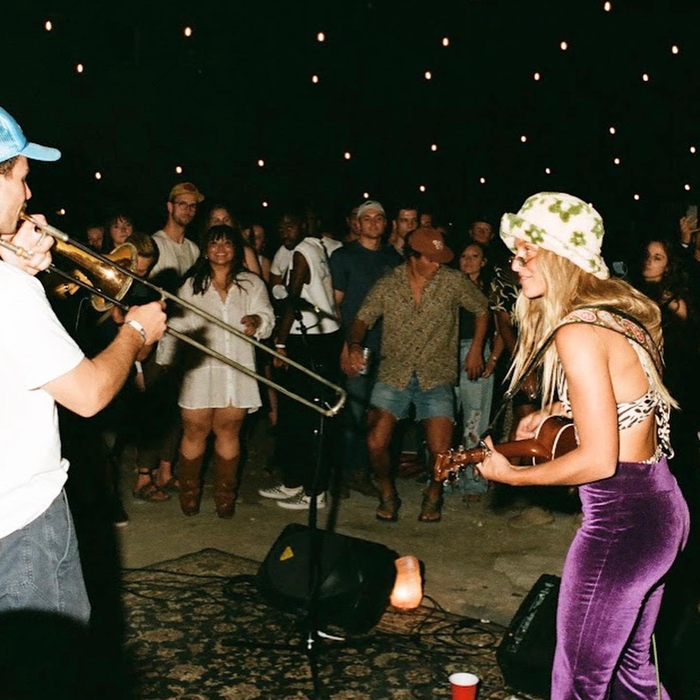 Musicians perform for a crowd under star-like lights, with a woman playing guitar and a man playing trumpet, audience watching.