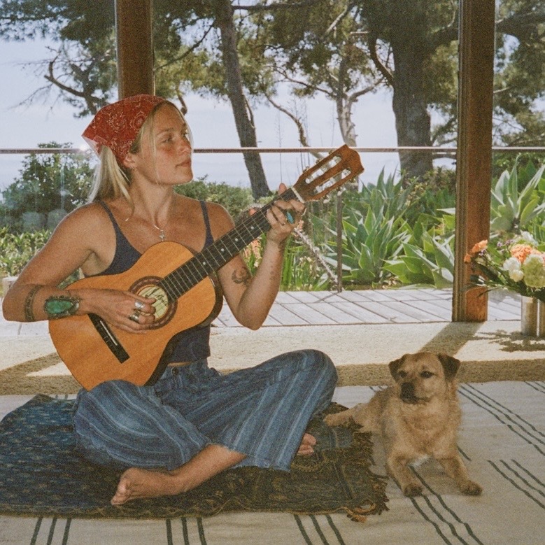 Woman sitting on a rug playing guitar with a dog beside her, indoors with large windows and greenery outside.