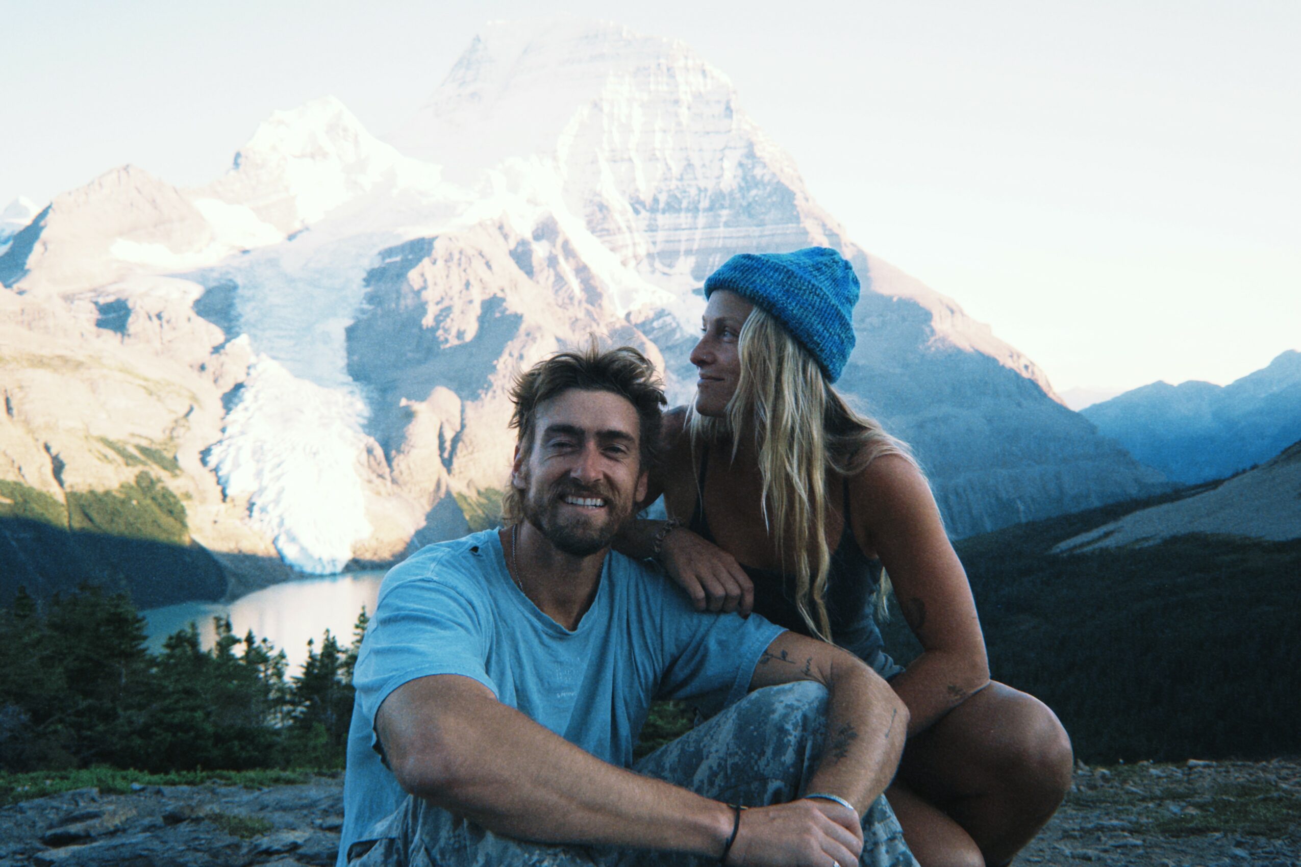 A smiling man and woman sit outdoors with mountains and a lake in the background.