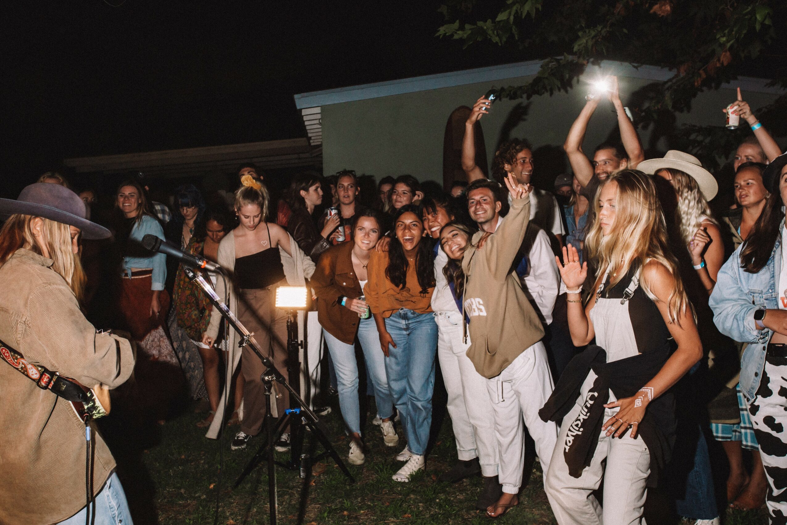 Group of people gathered outdoors at night, some dancing and taking photos, with a musician playing guitar in the foreground.