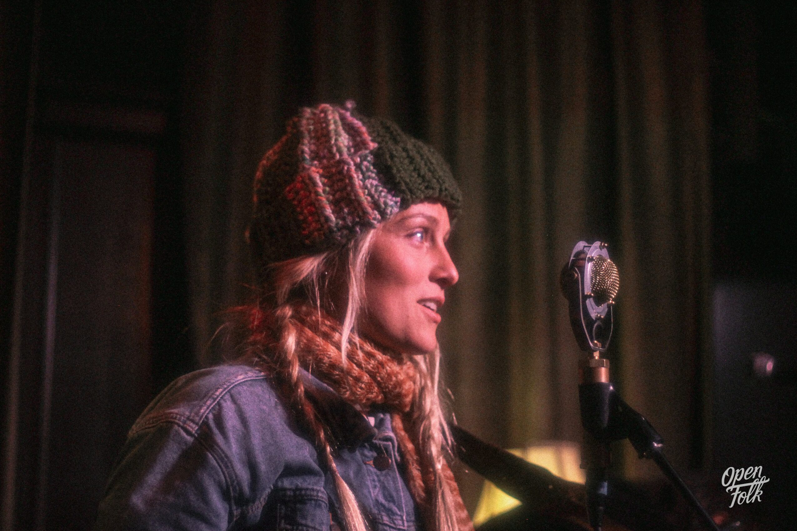 Woman wearing a knit hat and denim jacket speaking into a microphone, with curtains in the background.