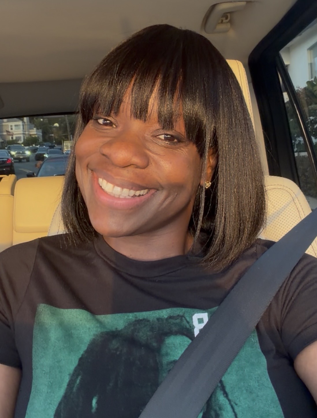 Smiling woman with shoulder-length dark hair and bangs, sitting in a car, wearing a black shirt, sunlight on face
